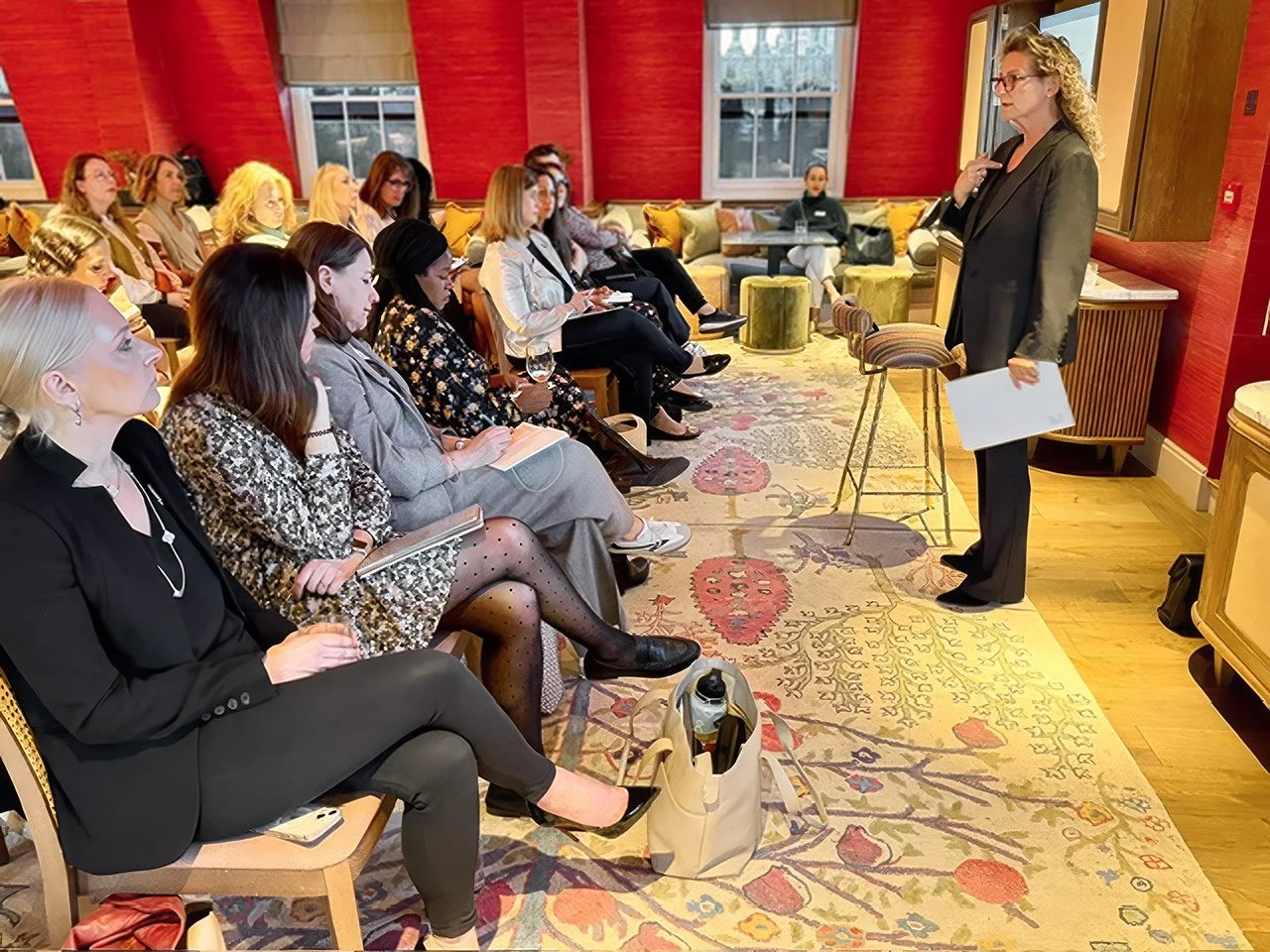 Deborah Dor giving a presentation to an audience of women seated in a conference room with red walls and large windows.