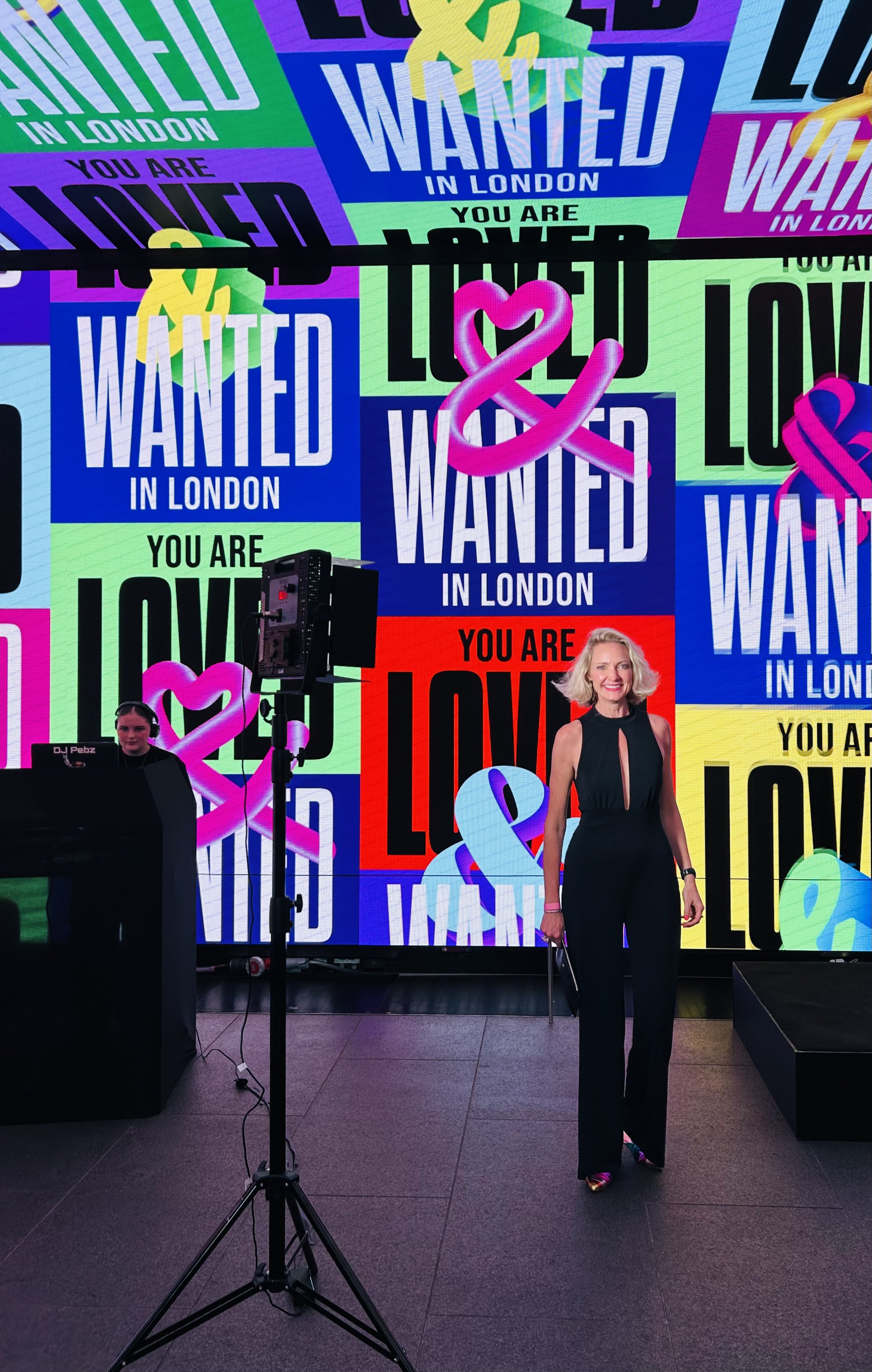 Heidi Mallace stands in front of a vibrant digital display with multiple 'Wanted in London' and 'You Are Loved' posters in various colours. She is smiling, dressed in black, and holding a small bag.