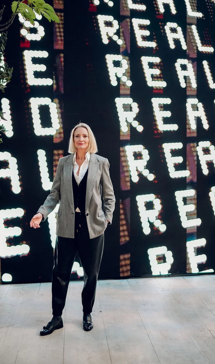Heidi Mallace in a grey blazer and black pants standing in front of a brightly lit digital sign that spells out words "REAL"