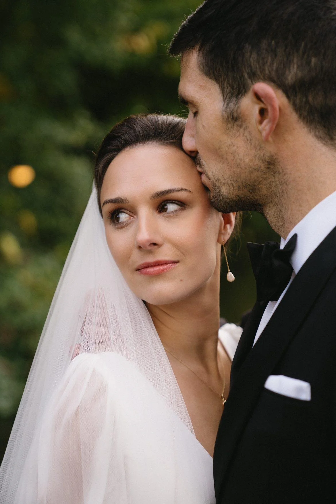 A bride and groom on their wedding day, with the groom gently kissing the bride's forehead against a blurred green outdoor background.