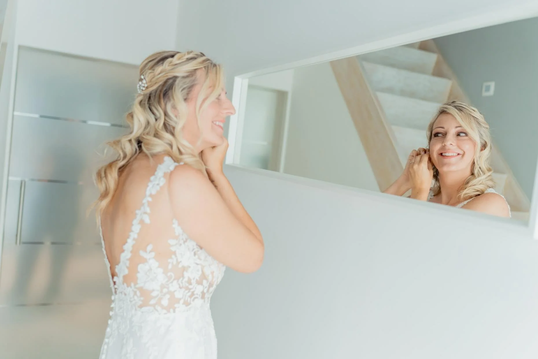 A woman in a white wedding dress looking at herself in a mirror and smiling.