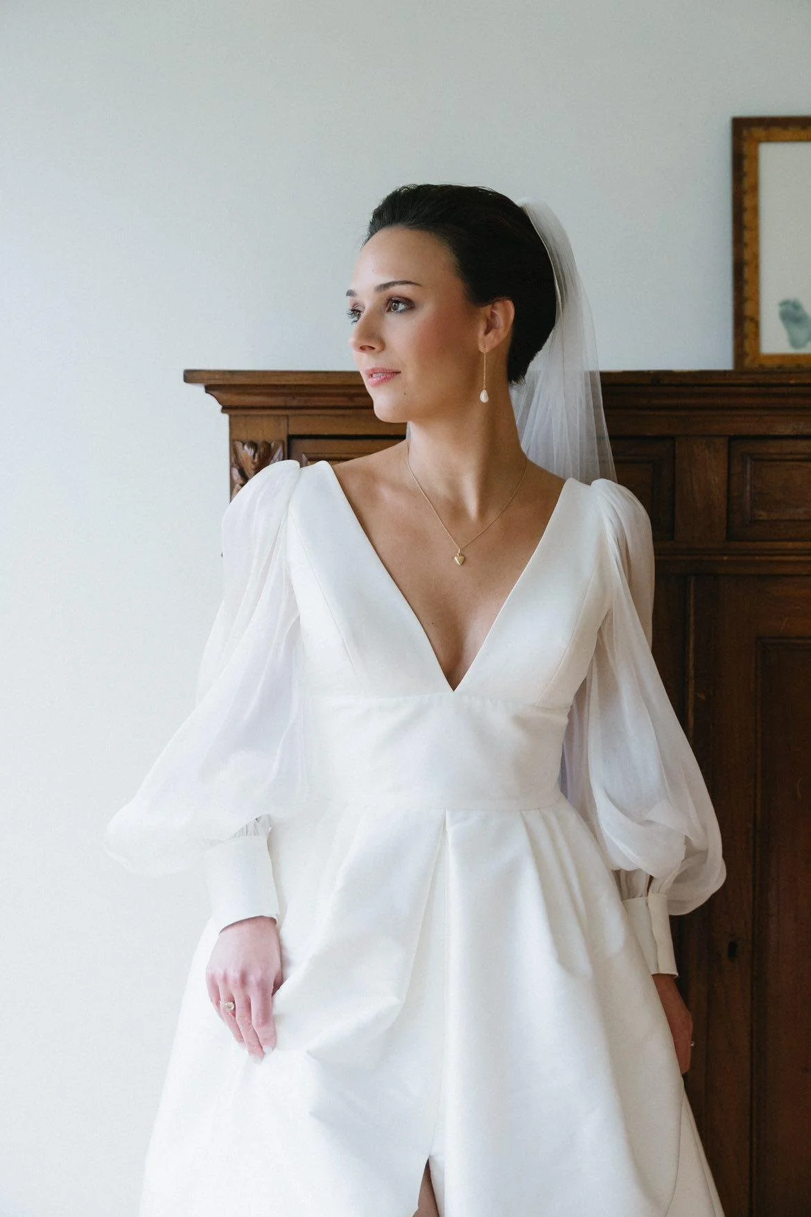 A bride with dark hair in an elegant white wedding dress, standing indoors near a wooden headboard or cabinet, looking to the side. She is wearing pearl earrings, a delicate necklace, and a veil.
