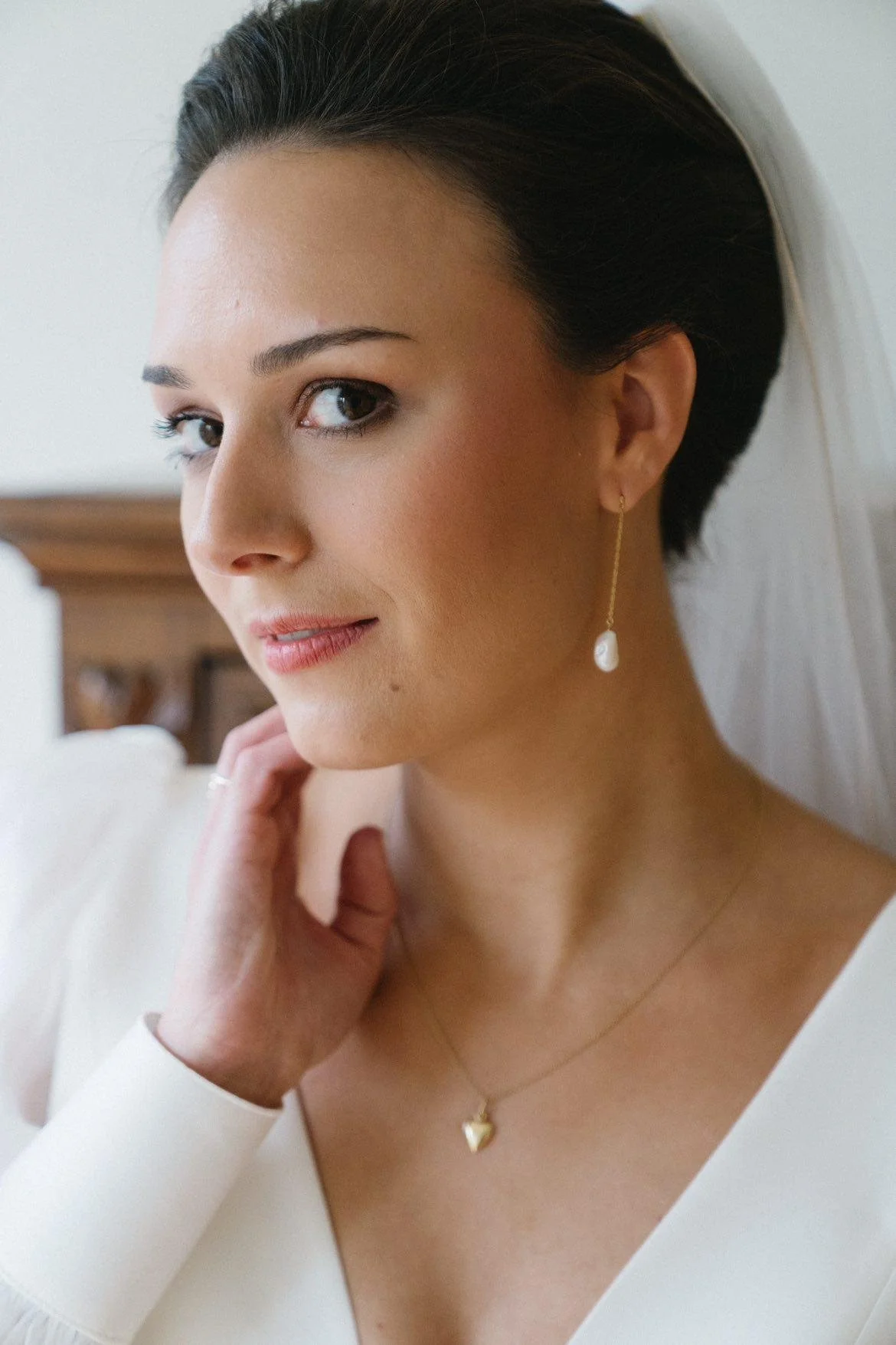 A woman with short dark hair wearing pearl drop earrings and a heart-shaped pendant necklace, dressed in a white outfit, standing indoors with a wooden piece of furniture in the background.