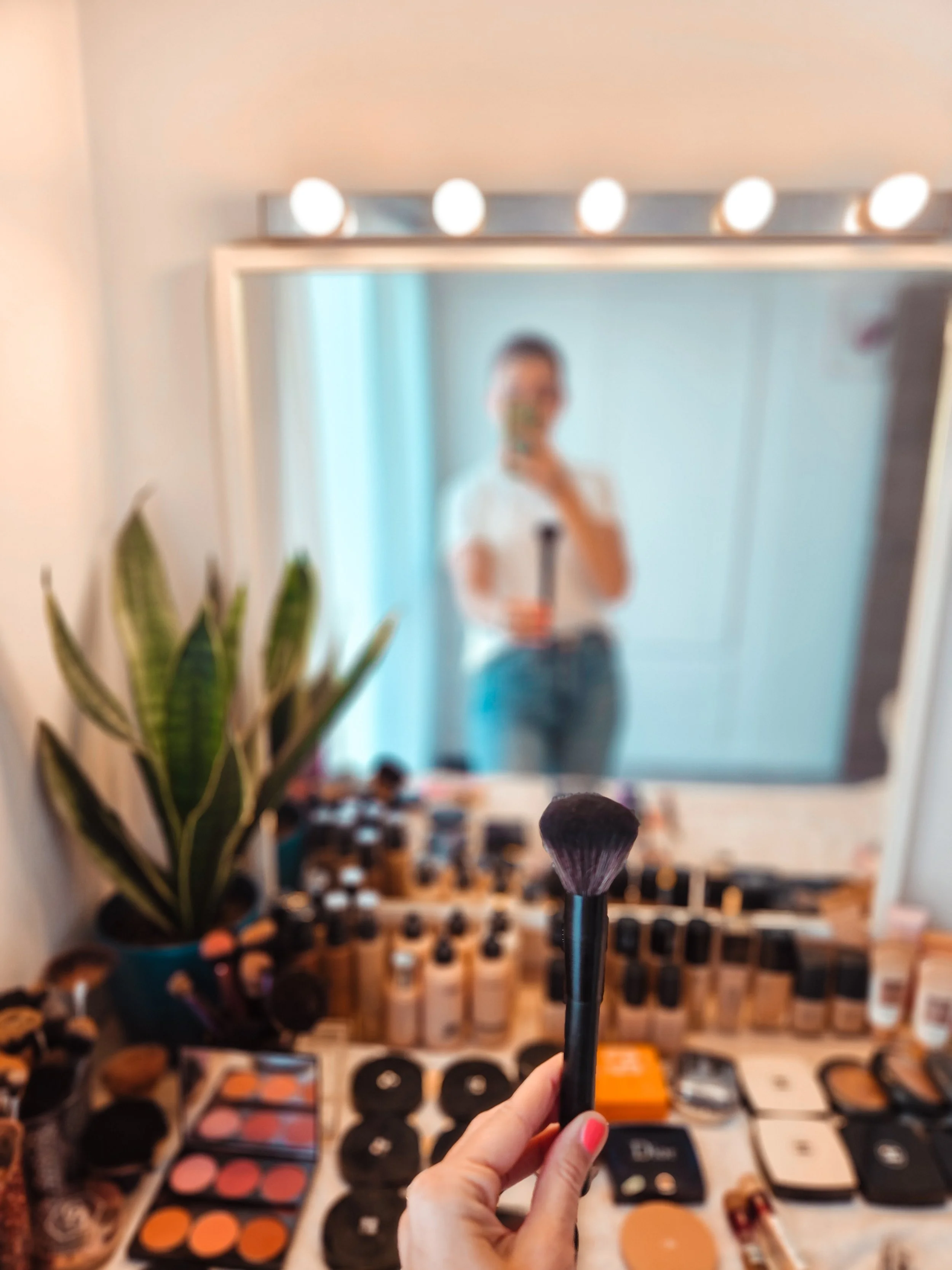 A hand holding a makeup brush in front of a mirror, with a woman taking a selfie outside the mirror, a makeup table with various cosmetics, and a potted plant in the background.