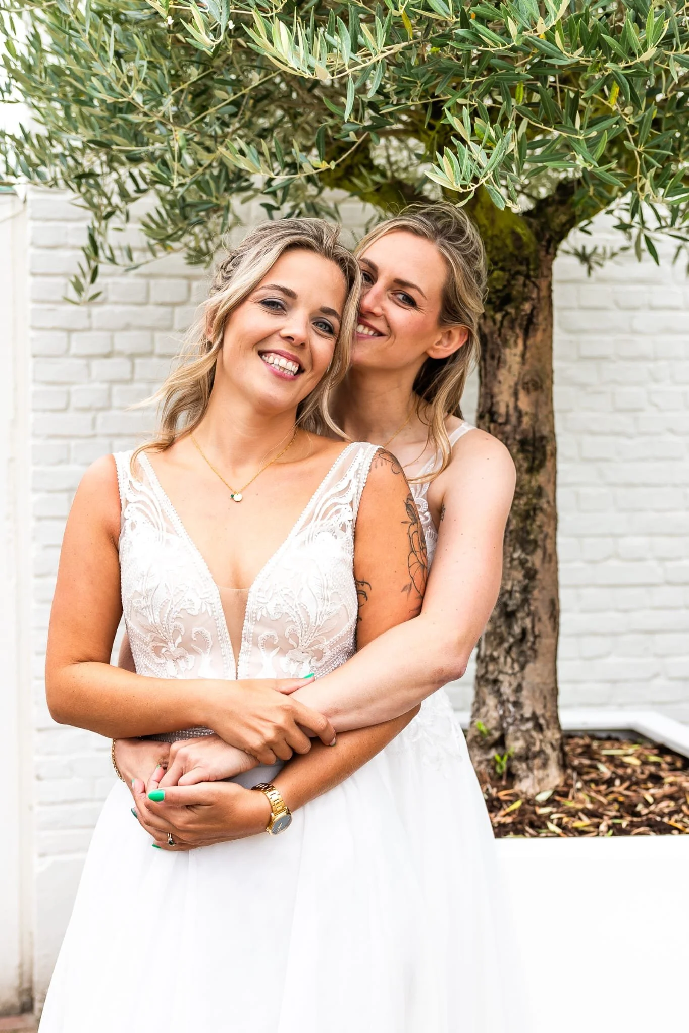 Two women in white dresses embracing in front of a white brick wall and a tree, smiling at the camera.