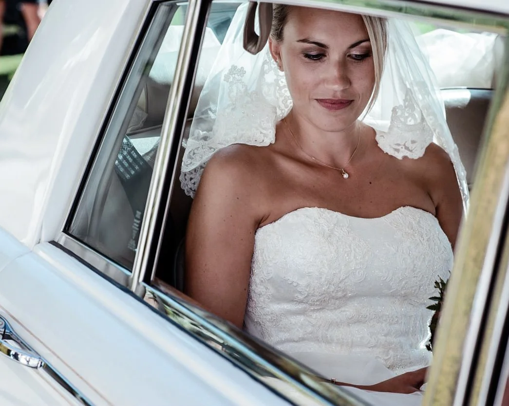 A bride wearing a white strapless wedding dress and lace veil sitting inside a vintage car, looking down with a serene expression.