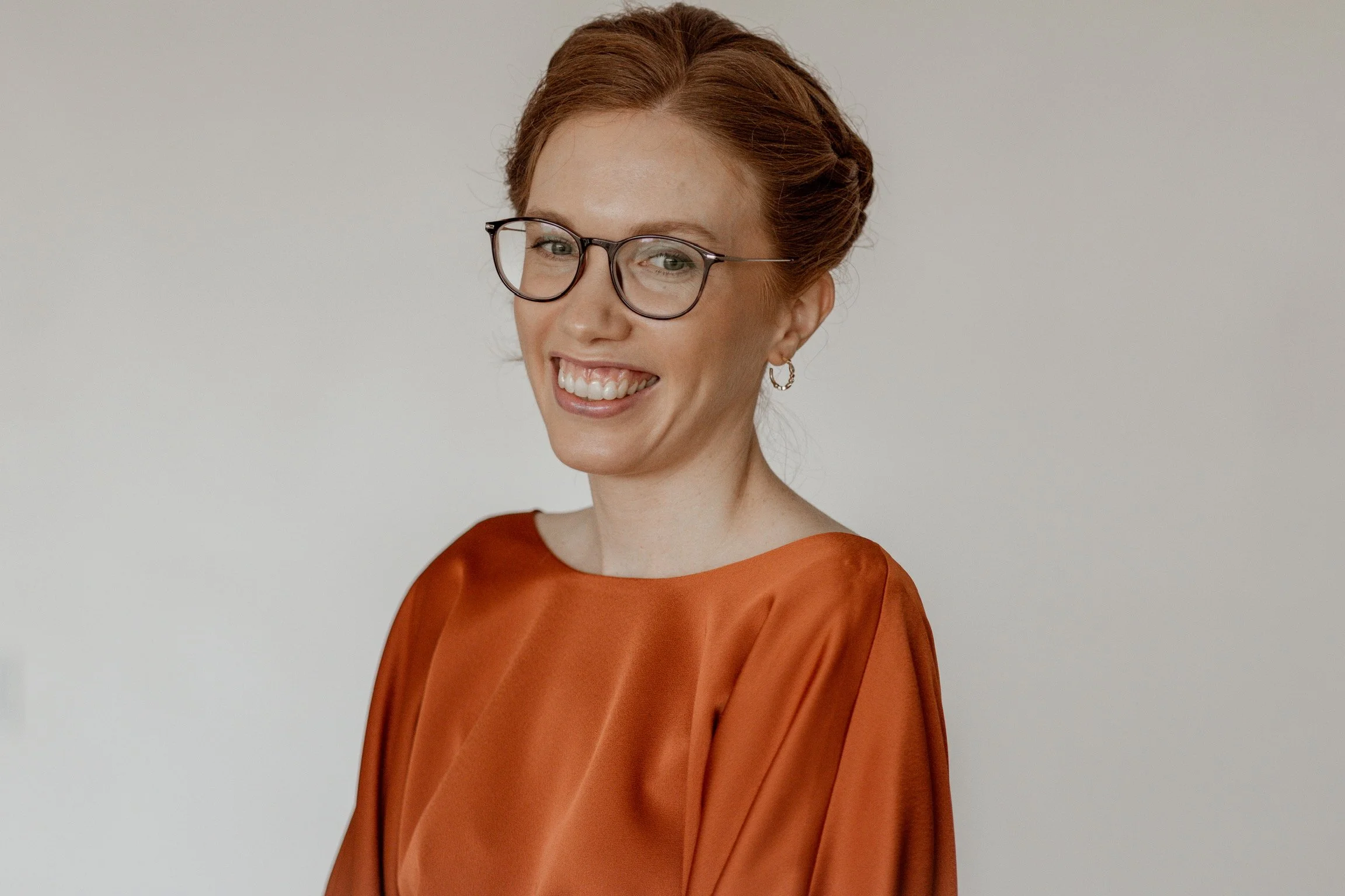 A woman with red hair, glasses, and hoop earrings smiling while wearing an orange satin blouse.