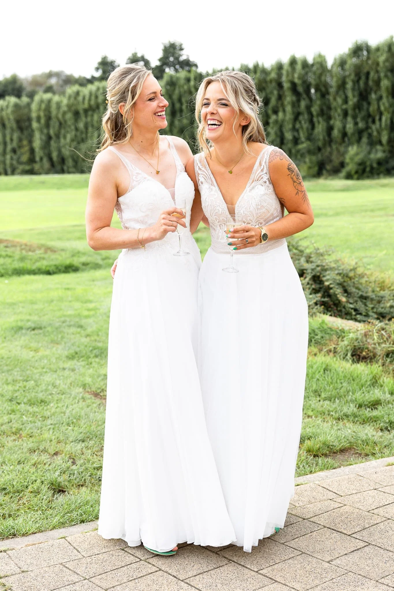 Two women in white dresses outdoors, smiling and holding champagne glasses, celebrating at a wedding or special event.