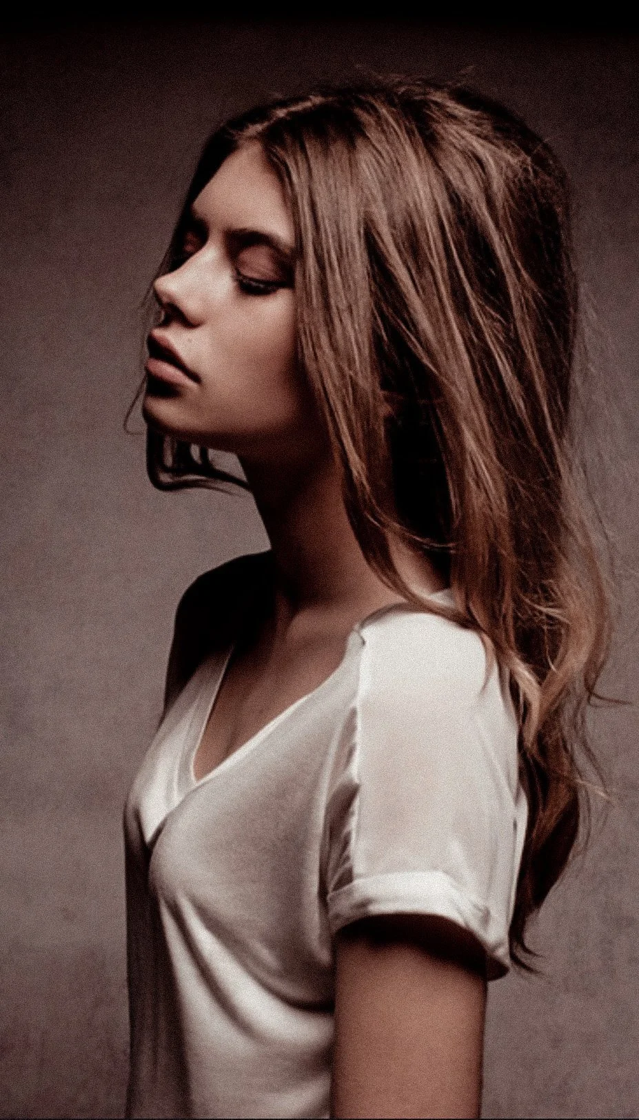 Side profile of a young woman with wavy brown hair, closed eyes, wearing a white top, against a neutral background.