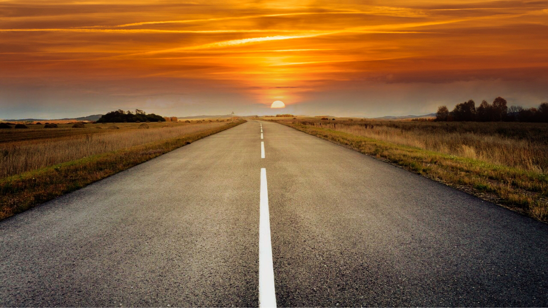 A straight empty road stretching into the horizon during sunset with an orange and yellow sky and clouds.