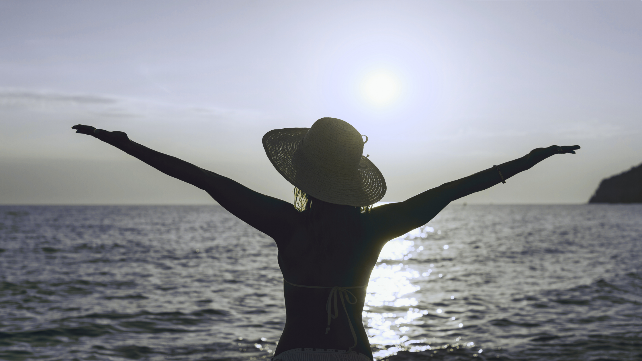 Silhouette of a woman wearing a large sun hat with arms outstretched on a beach at sunset.