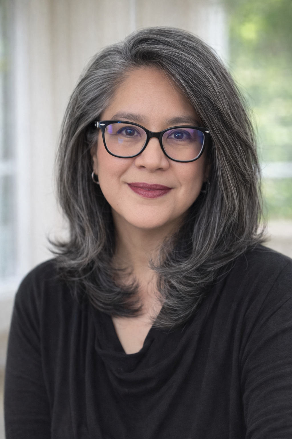 A woman with shoulder-length gray hair, wearing black glasses and a black top, smiling at the camera. Background shows a window with green trees outside.