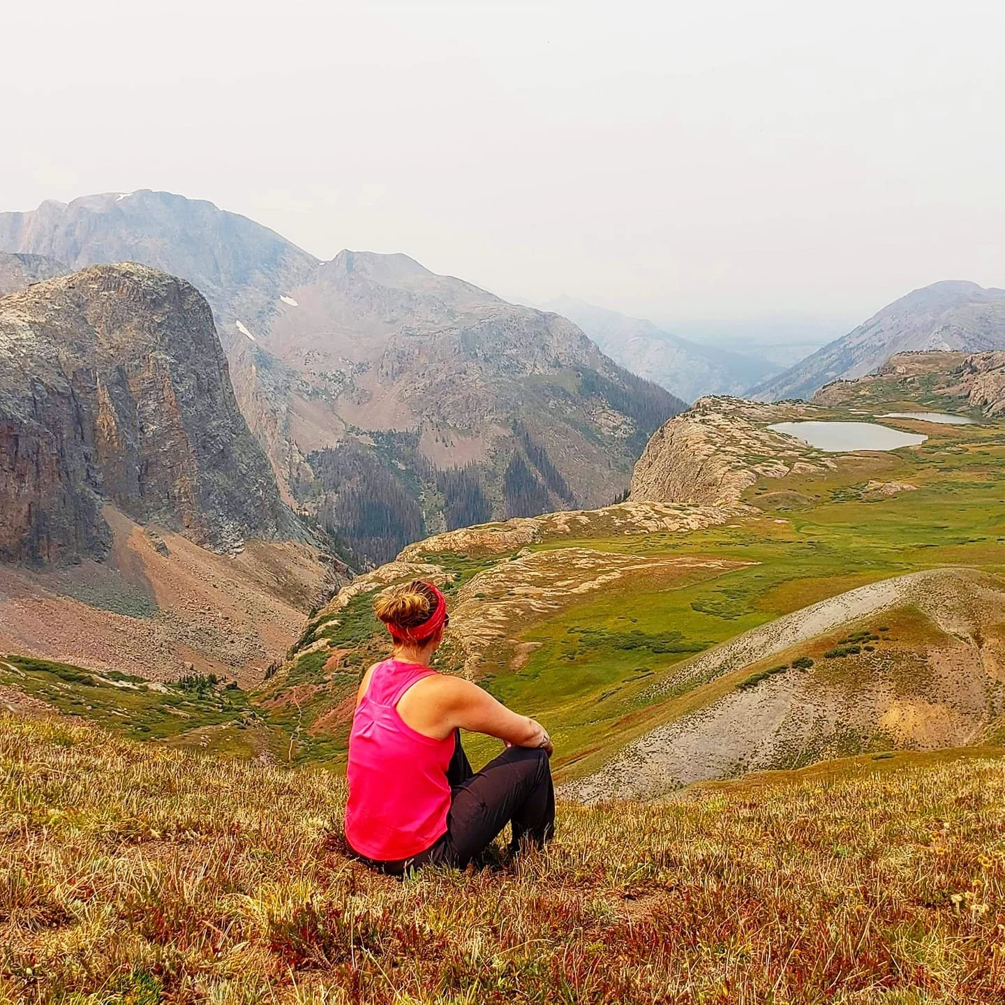 A woman sitting on grassy terrain in a mountainous landscape, wearing a pink tank top and a red headband, looking at distant mountains and a lake.