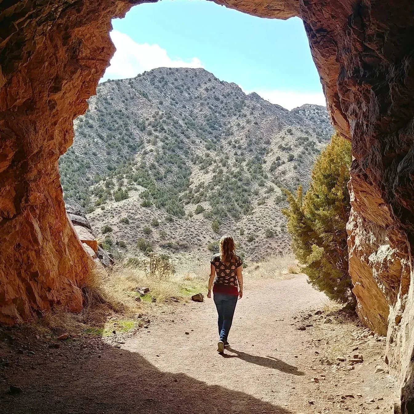 A woman with long hair walking towards an outdoor mountain view through a rocky cave opening, with dry grass and trees surrounding the trail.