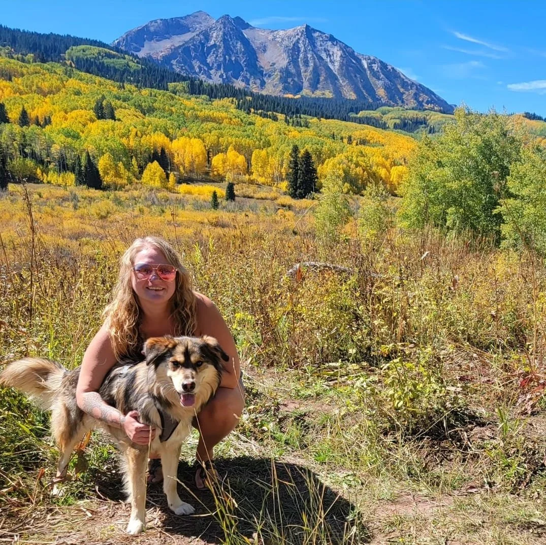 A woman with long blonde hair and pink sunglasses crouching next to a fluffy dog with tan, black, and white fur in a field of tall grass, with a mountain range and colorful autumn trees in the background on a bright, clear day.