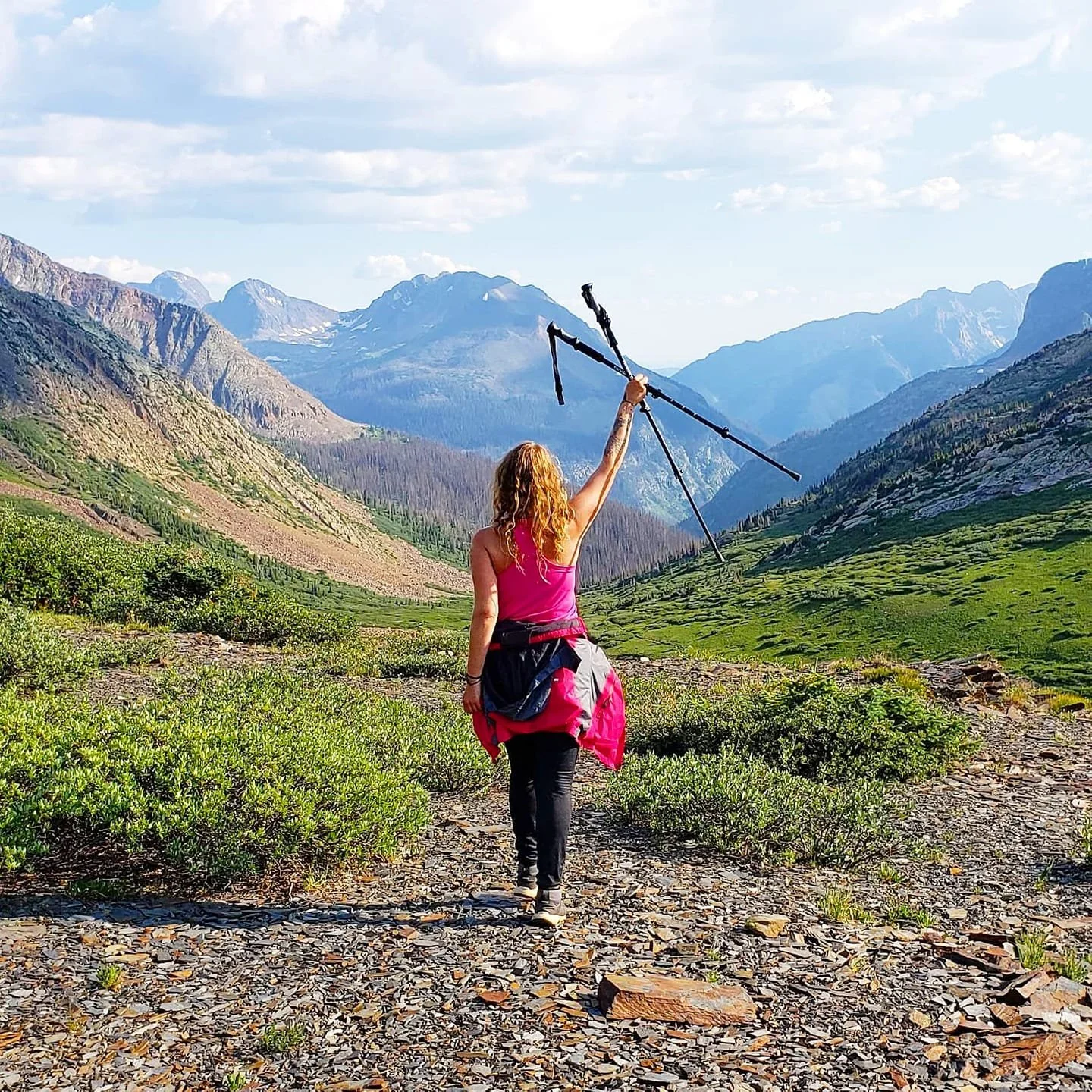 A woman walking through a scenic mountain valley holding trekking poles overhead.
