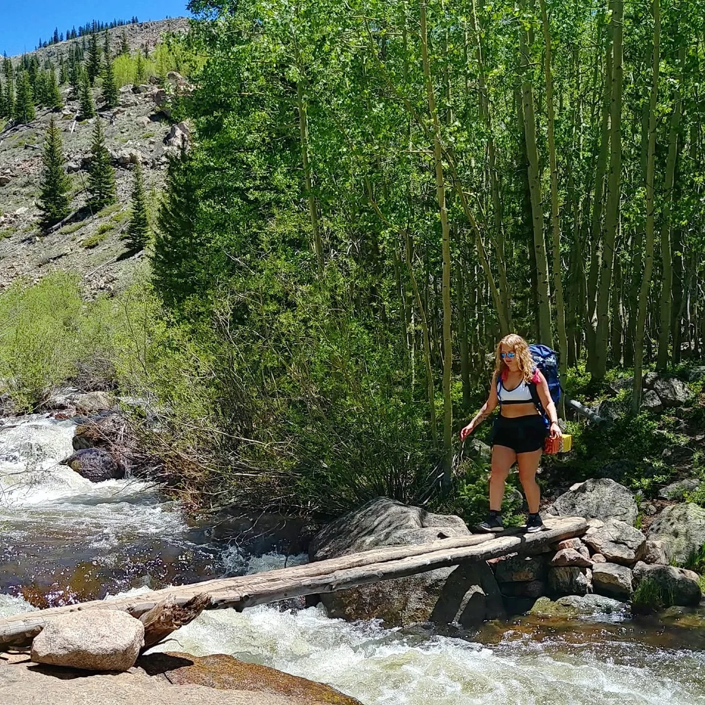 A woman with curly blonde hair, wearing sunglasses, a sports bra, and shorts, crosses a small wooden bridge over a rushing river in a forested mountain area with green trees and rocky terrain.