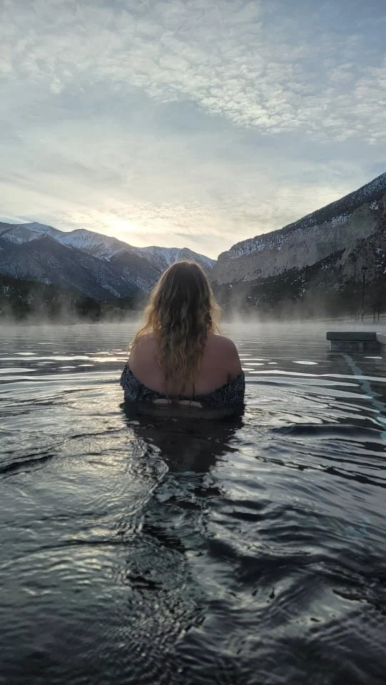 A person with long hair, viewed from behind, sitting in a hot spring or pool with mountains in the background, mist rising from the water, and a cloudy sky overhead.