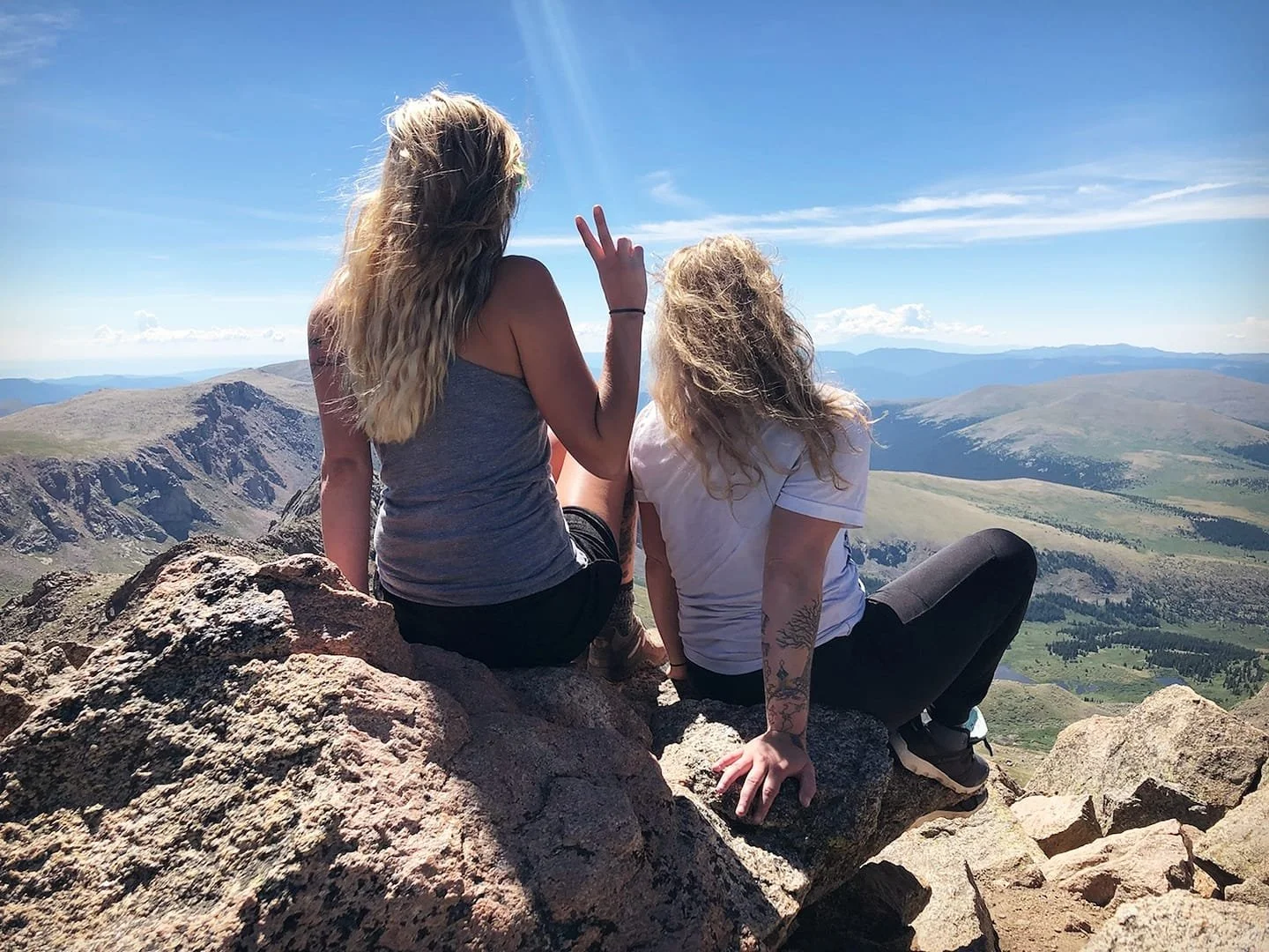 Two women sitting on rocks on a mountain peak, overlooking a scenic landscape of rolling hills, valleys, and distant mountains, under a bright blue sky with some clouds.