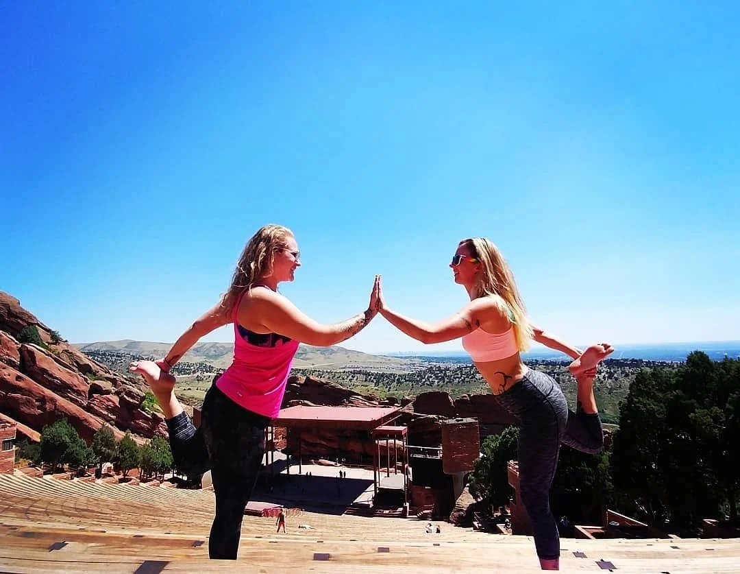 Two women practicing yoga outdoors on a wooden platform, balancing on one leg with hands touching in a yoga pose against a landscape with rocks, trees, and a clear blue sky.