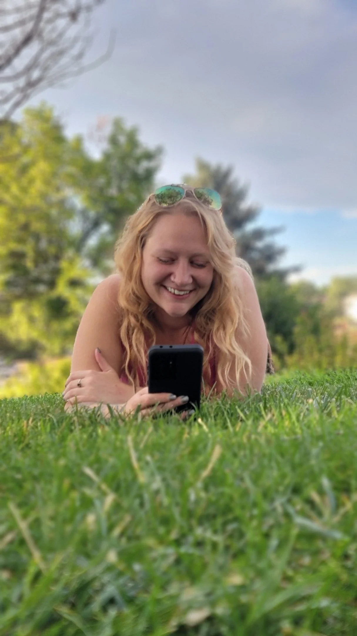 Woman with long blonde hair, smiling, lying on grass, looking at her phone, wearing sunglasses on her head, outdoors with trees and cloudy sky in the background.