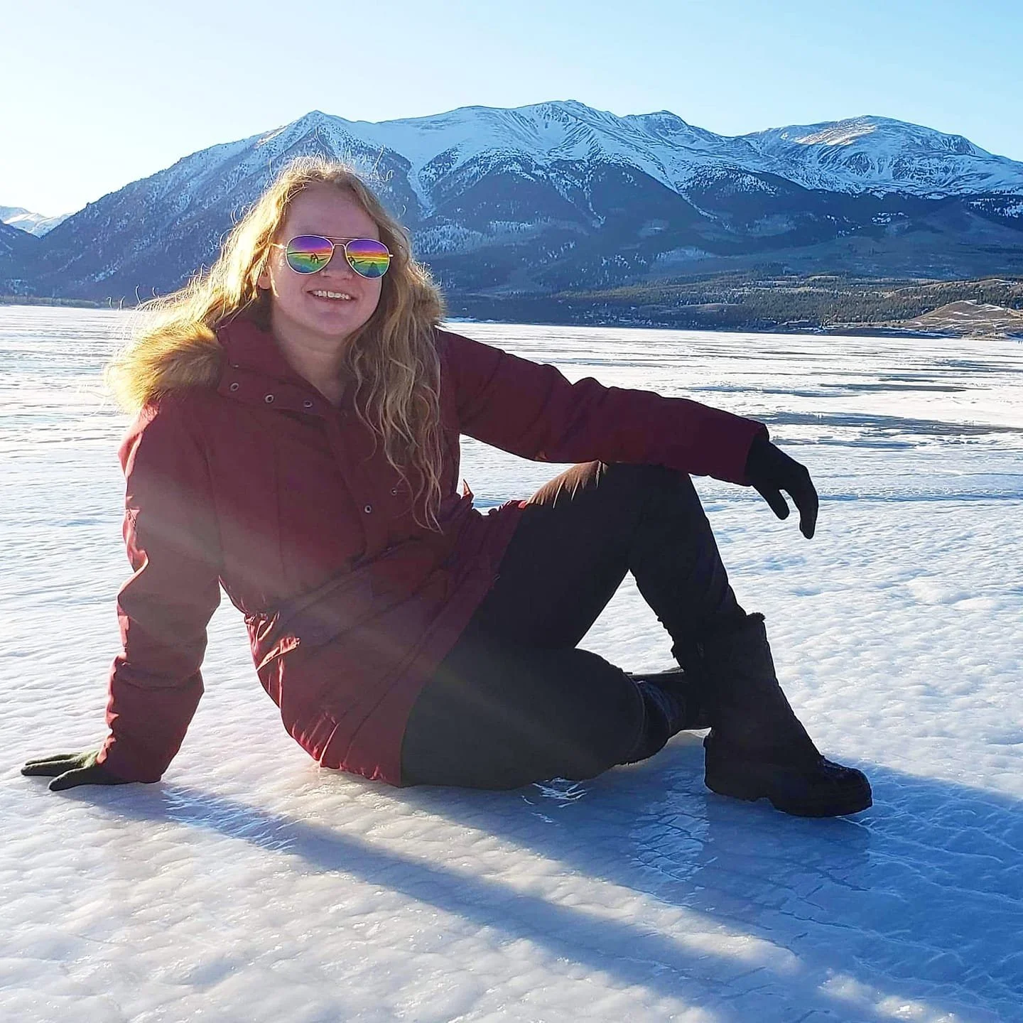 Woman sitting on snow-covered ground with mountains in the background, wearing sunglasses, a maroon winter coat with fur-lined hood, black gloves, black pants, and black boots, smiling.