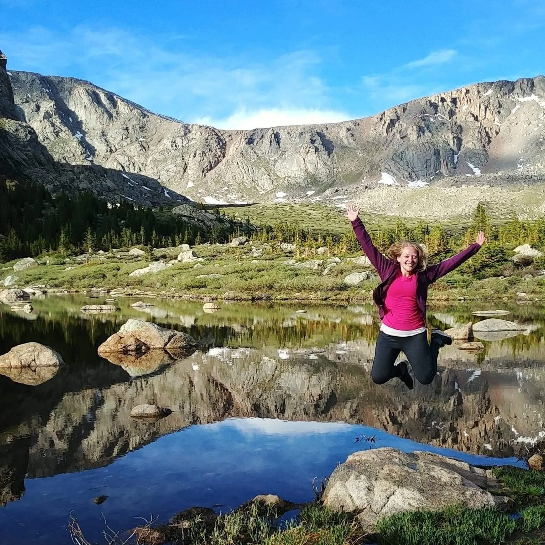 A woman jumping joyfully near a mountain lake with rocky and forested landscape in the background.