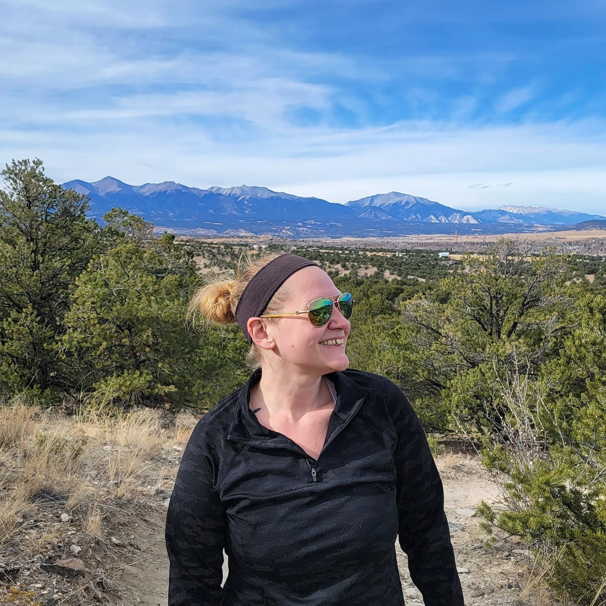 Woman with blonde hair in a ponytail, wearing sunglasses, a black headband, and a black zip-up athletic shirt, smiling and standing on a trail in a mountainous landscape with green trees and distant mountain range.