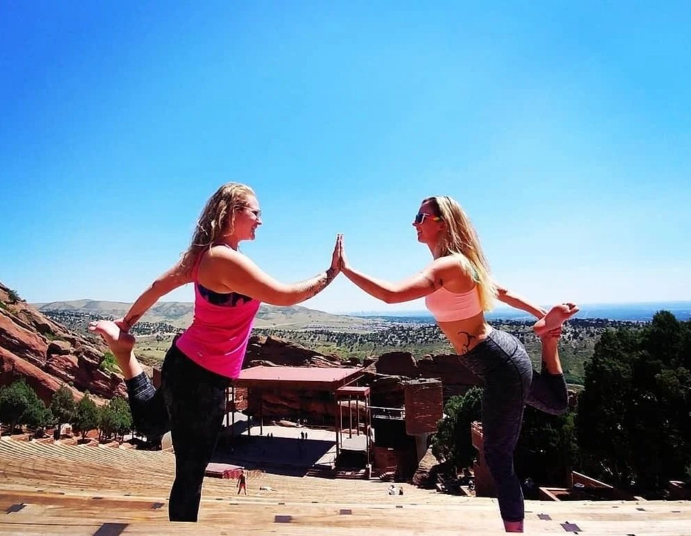 Two women practicing yoga outdoors on a clear sunny day, standing on one leg and touching hands in a balancing pose, with a scenic landscape in the background.