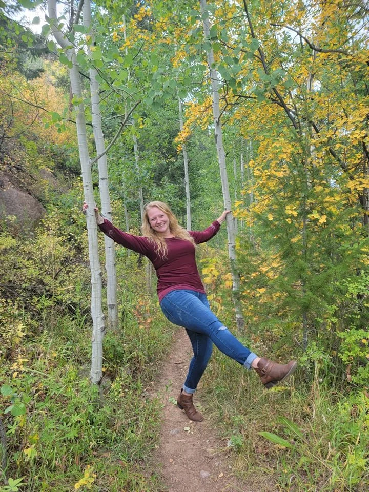 A woman with long blonde hair wearing a maroon sweater, blue jeans, and brown boots holding onto two white-barked trees, standing on a forest trail surrounded by green and yellow autumn leaves.