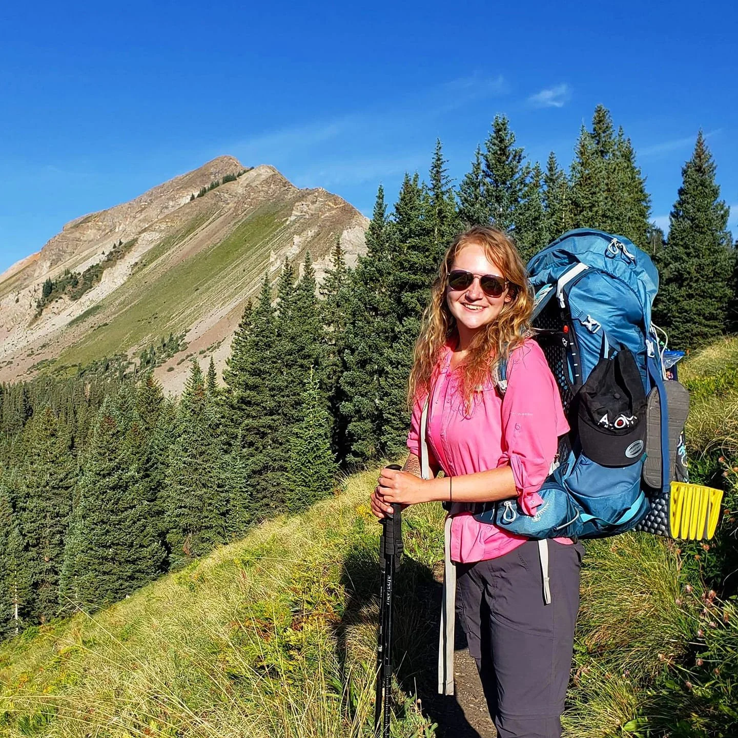 A woman hiking on a mountain trail with a large blue backpack, wearing sunglasses and a pink shirt, surrounded by tall green trees and mountain scenery under a clear blue sky.