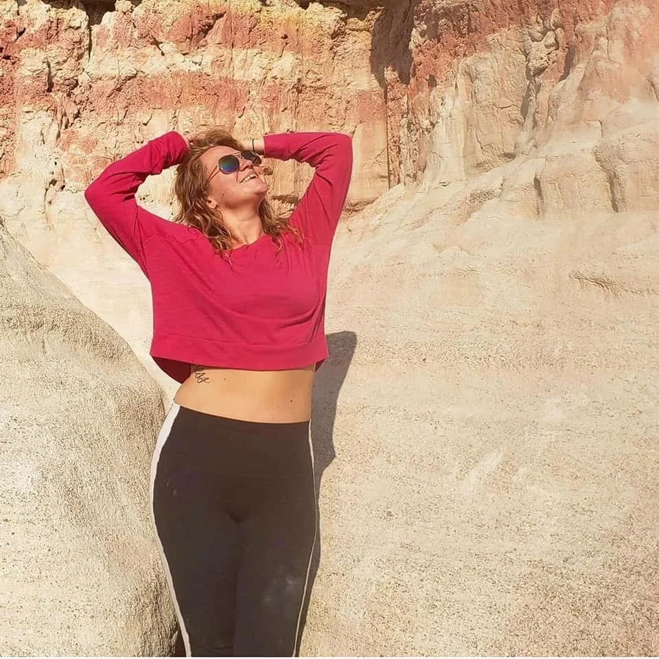 A woman smiling and posing with hands behind her head in a desert landscape with rocky cliffs in the background.