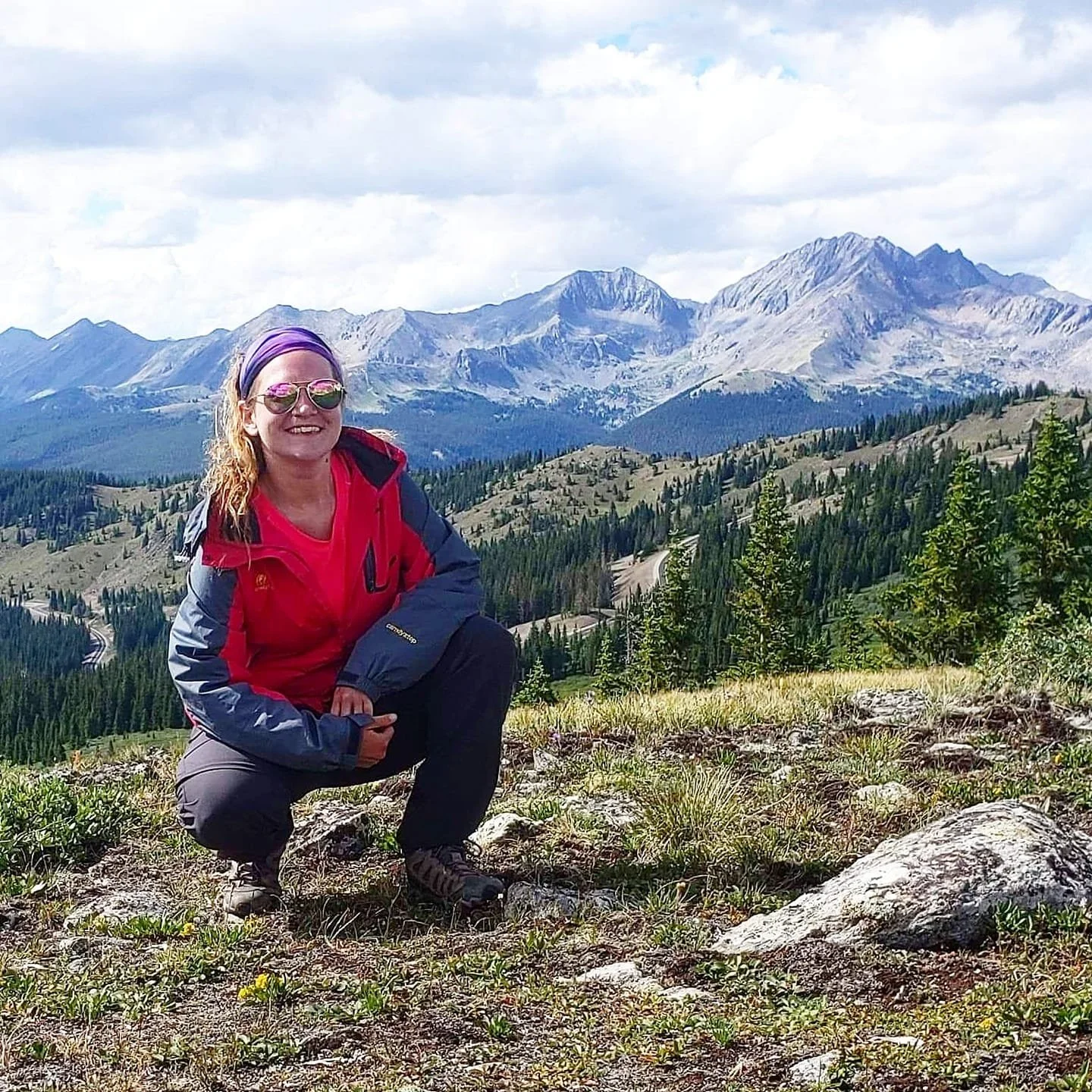Smiling woman in colorful outdoor jacket and sunglasses squatting on rocky terrain with mountains, trees, and cloudy sky in the background.