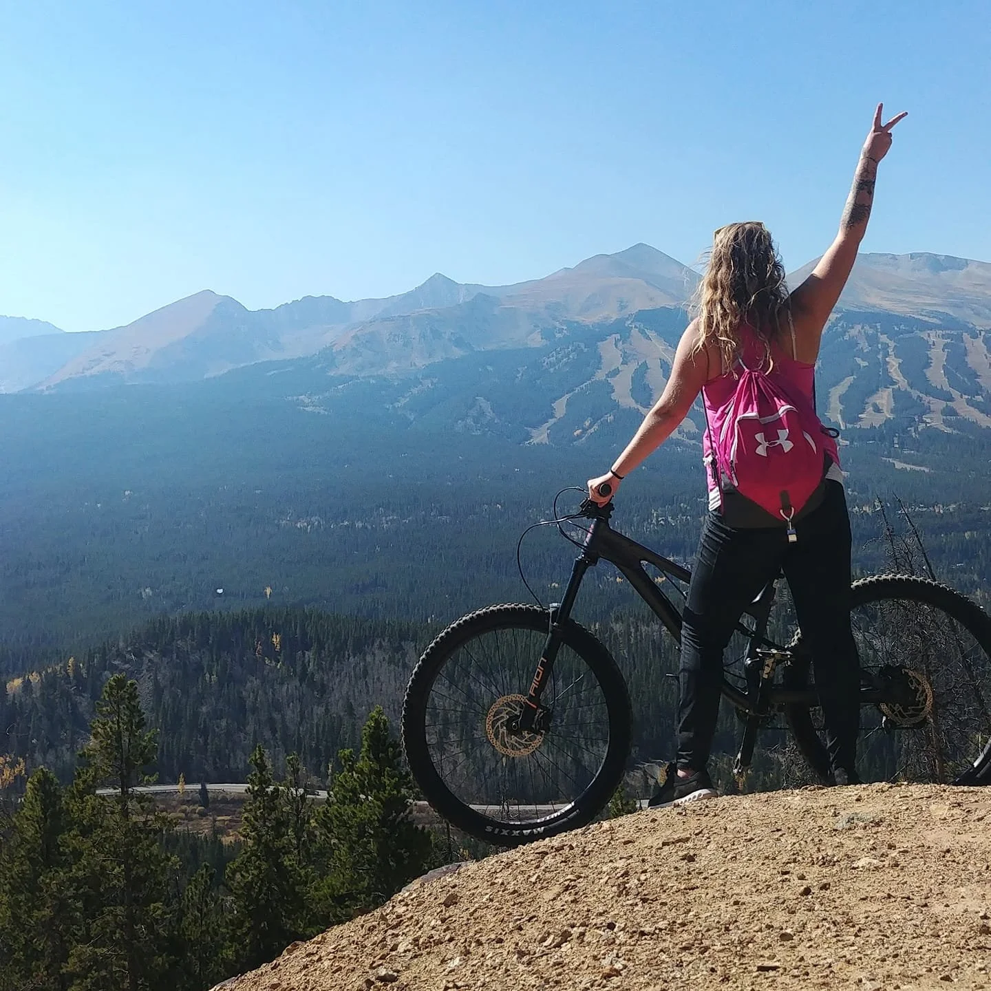 A woman with curly blonde hair standing beside a black mountain bike on a dirt hilltop, wearing a pink tank top and black pants with a pink backpack, raising one arm with a peace sign, overlooking a mountainous forested landscape under a clear blue sky.