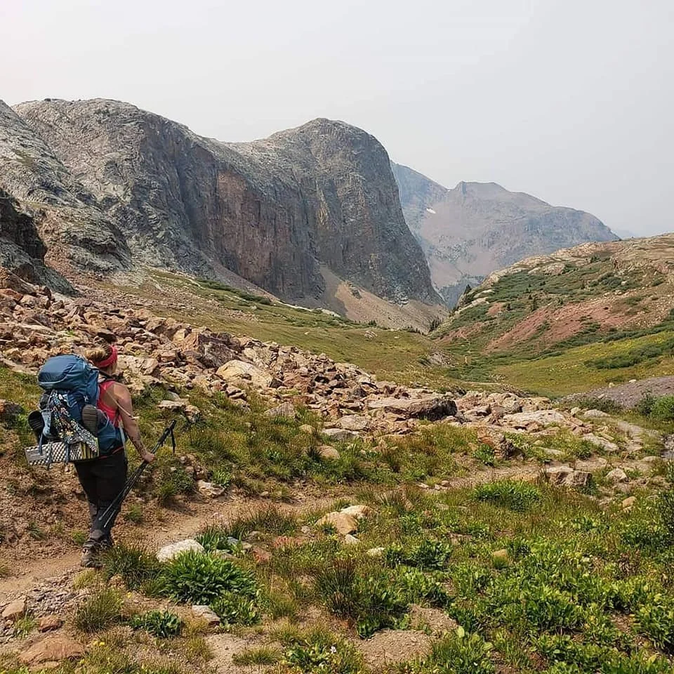 A hiker carrying a large backpack and trekking poles walking on a rocky trail through a mountainous landscape with green vegetation and large cliffs in the background.