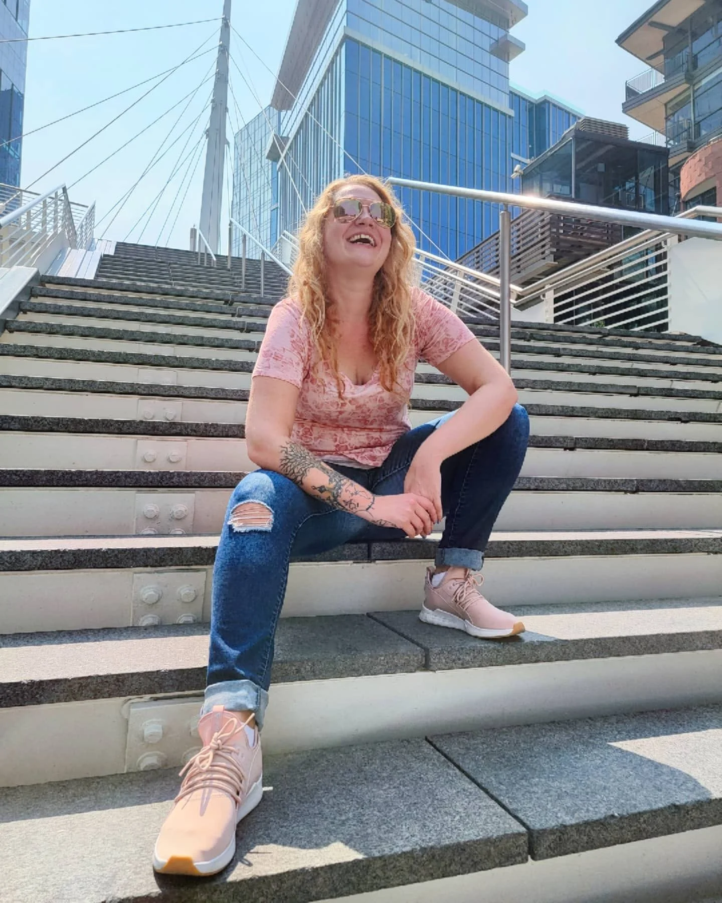 A woman with long red hair, wearing sunglasses, a pink shirt, ripped jeans, and pink sneakers, sitting on outdoor stairs with city buildings in the background, smiling and looking up.