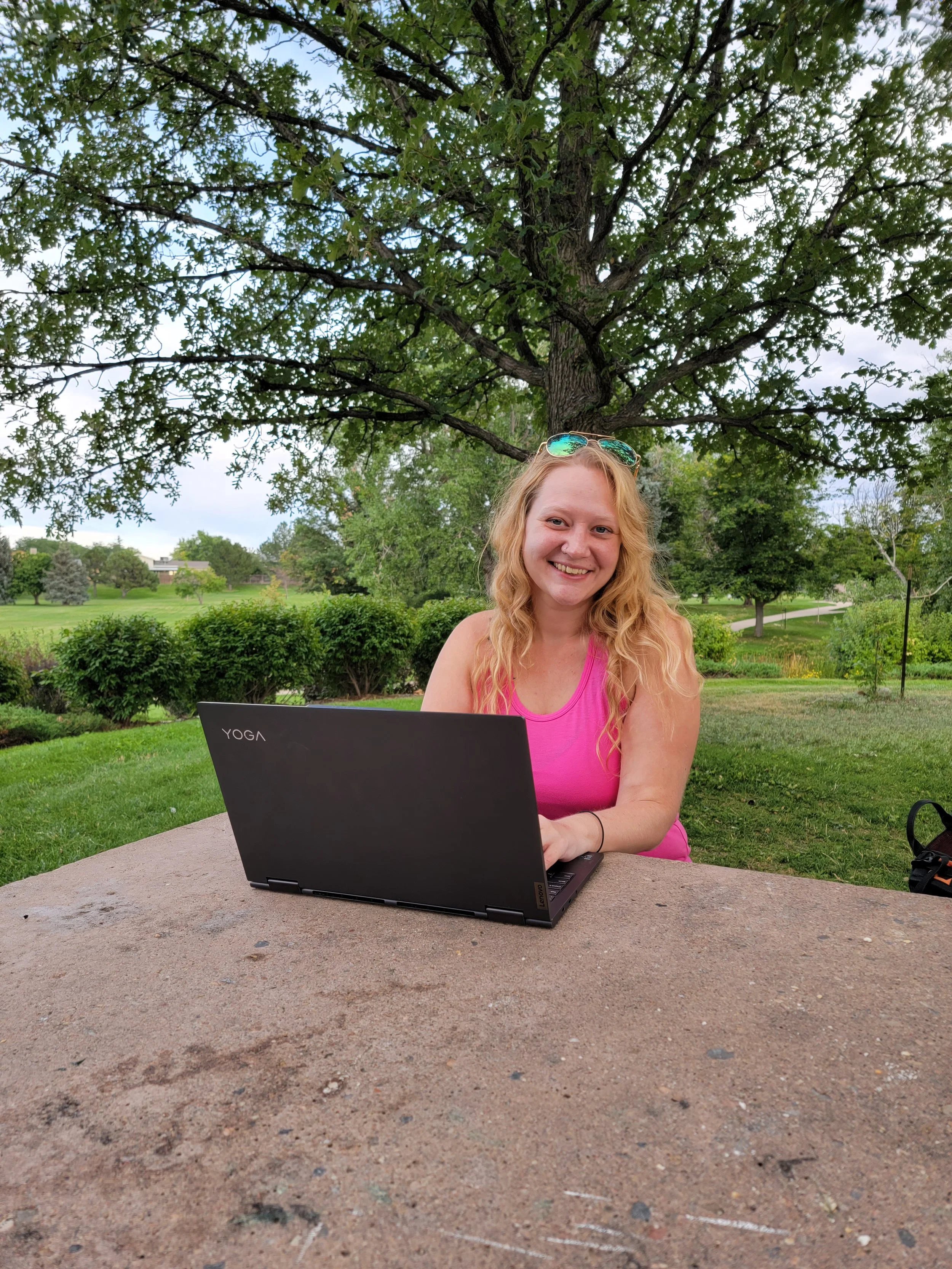 A woman with long, curly blonde hair sitting outside at a table with a black Lenovo Yoga laptop, smiling, wearing a pink sleeveless top, with sunglasses on her head, surrounded by lush green trees and grass on a cloudy day.