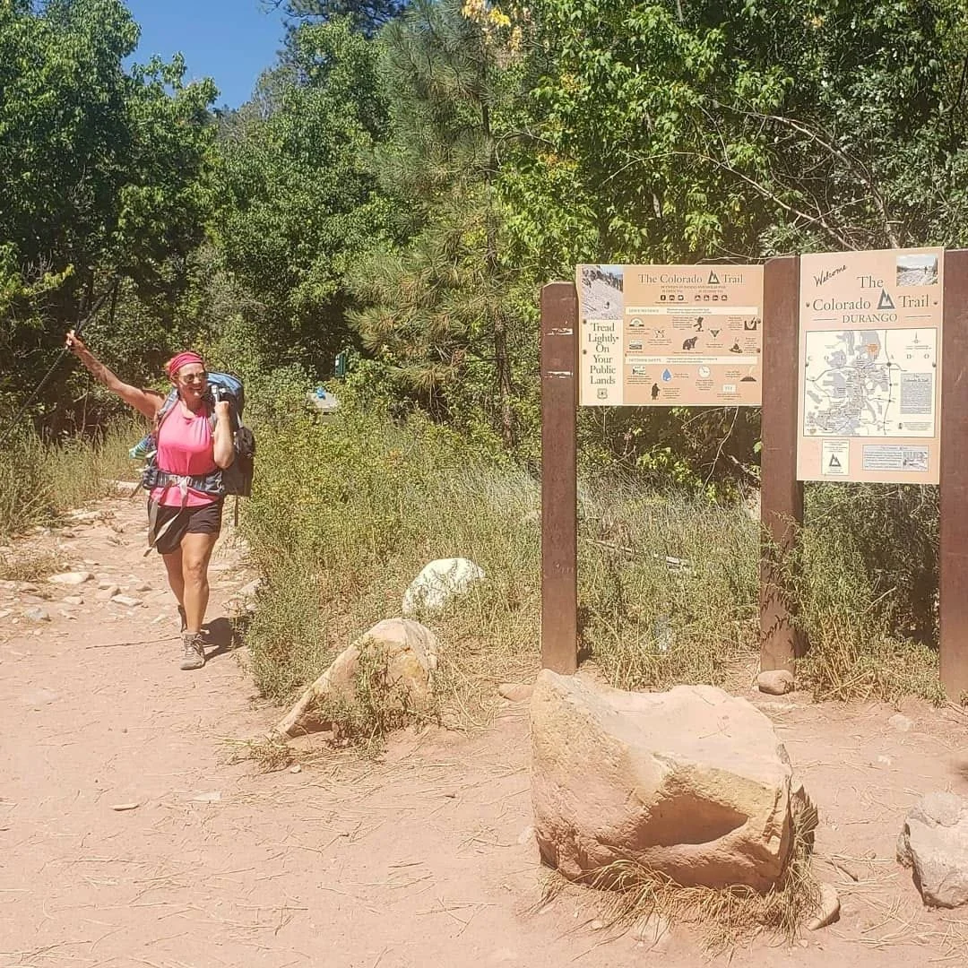 A woman with a pink top and black shorts hiking on the Colorado Trail near a trailhead sign with green trees in the background.