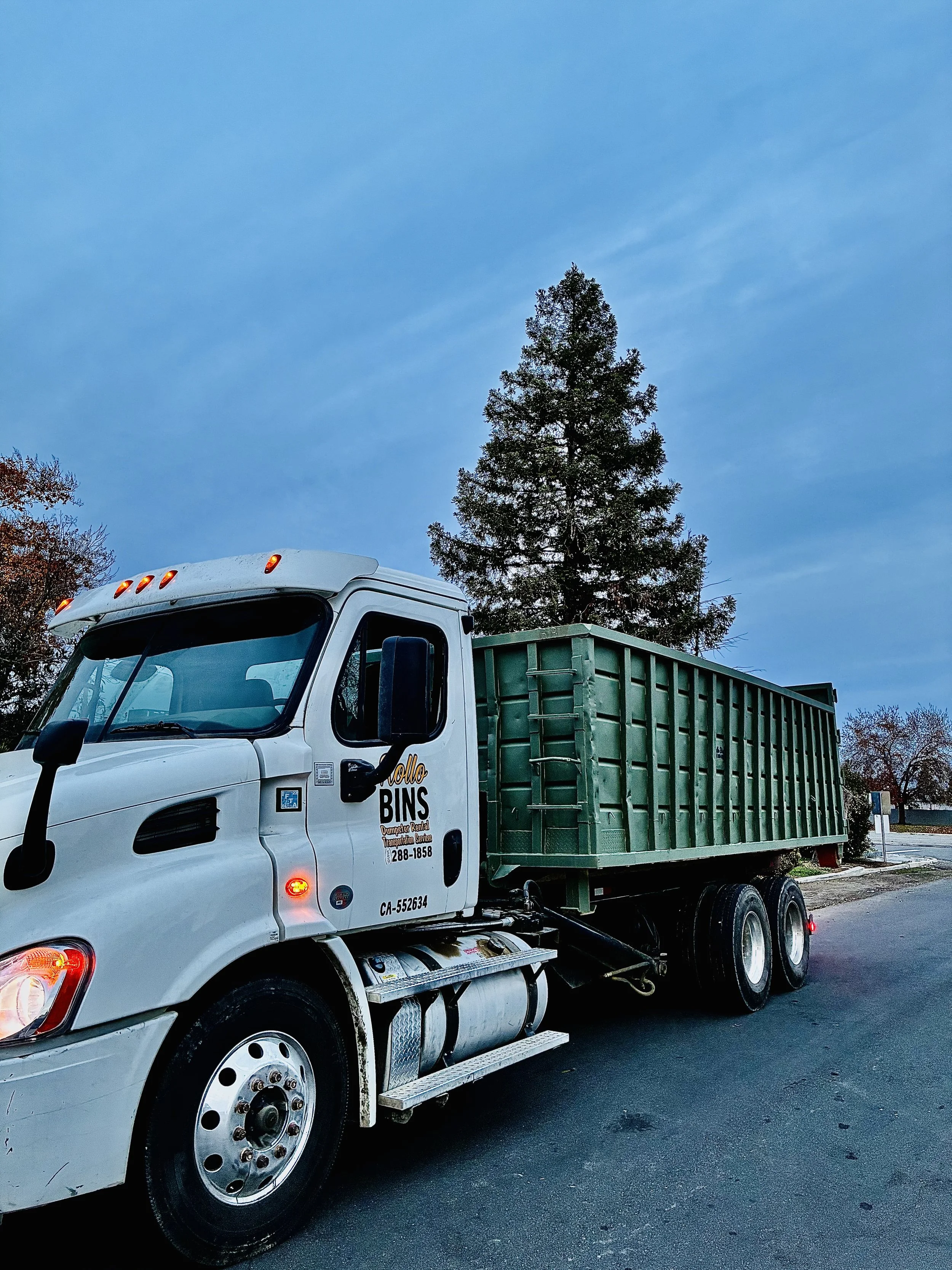 A white garbage truck with green dumpster container parked on the street under a blue sky, with trees in the background.