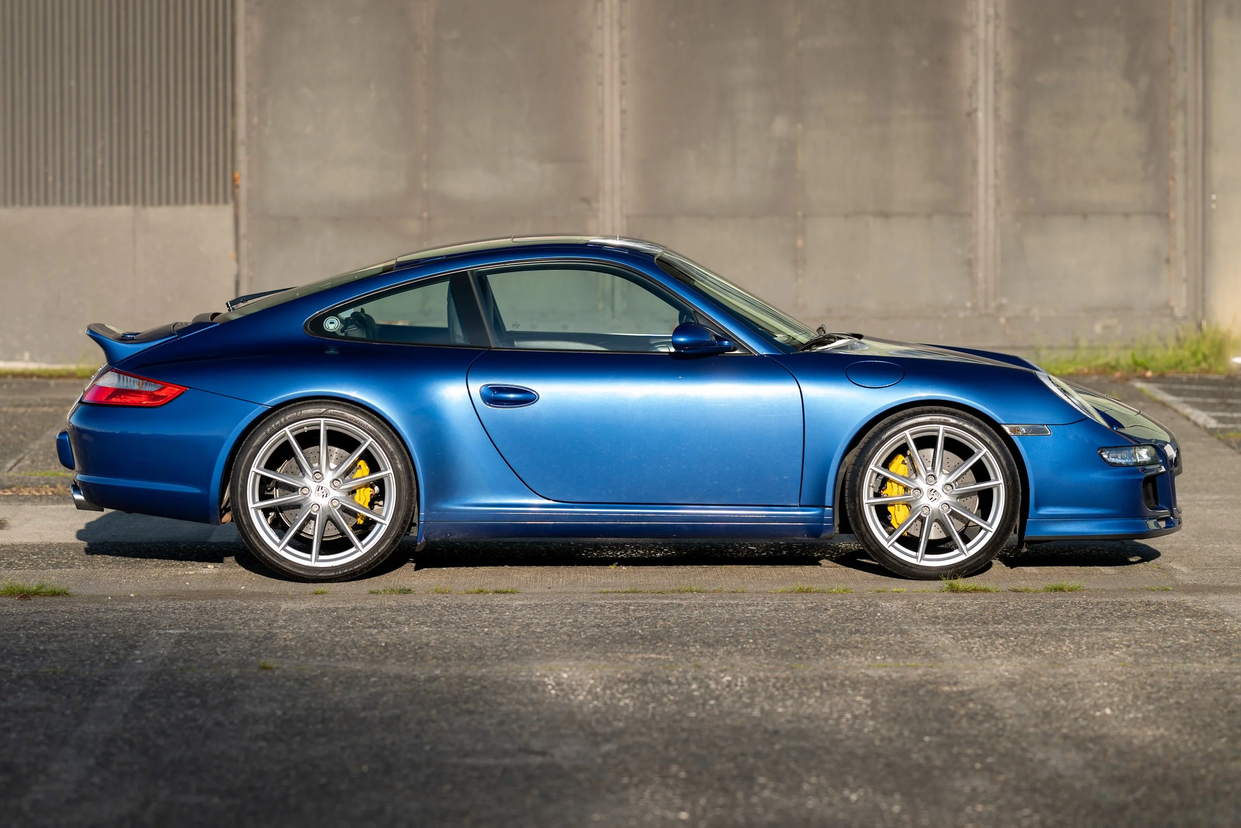 A blue sports car parked on a paved surface with a gray concrete wall in the background.