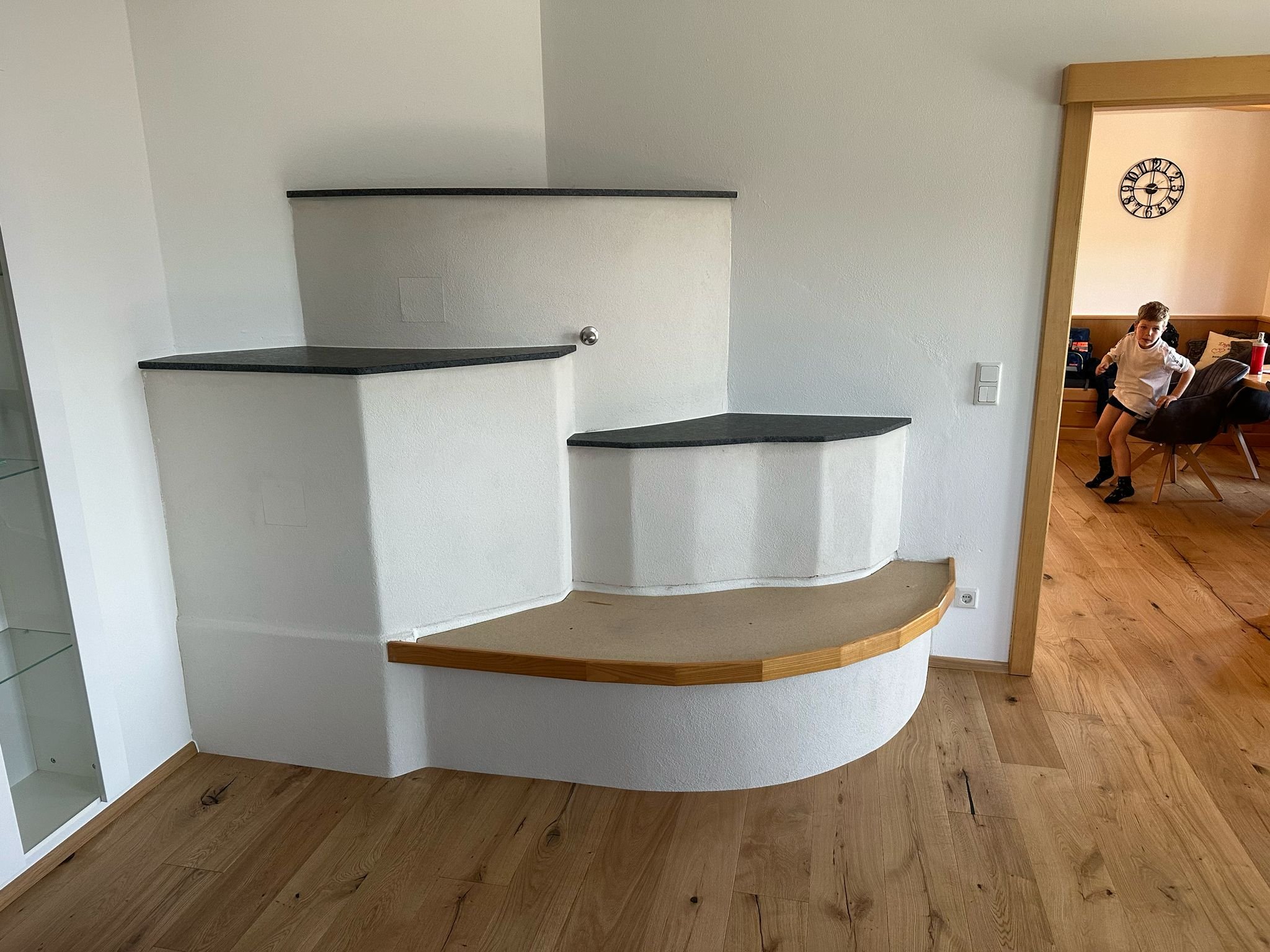 Ein weißer, mehrstufiger, halbrunder, modernisierter Kachelofen mit schwarzen Oberflächen und einer hölzernen Ofenbank.