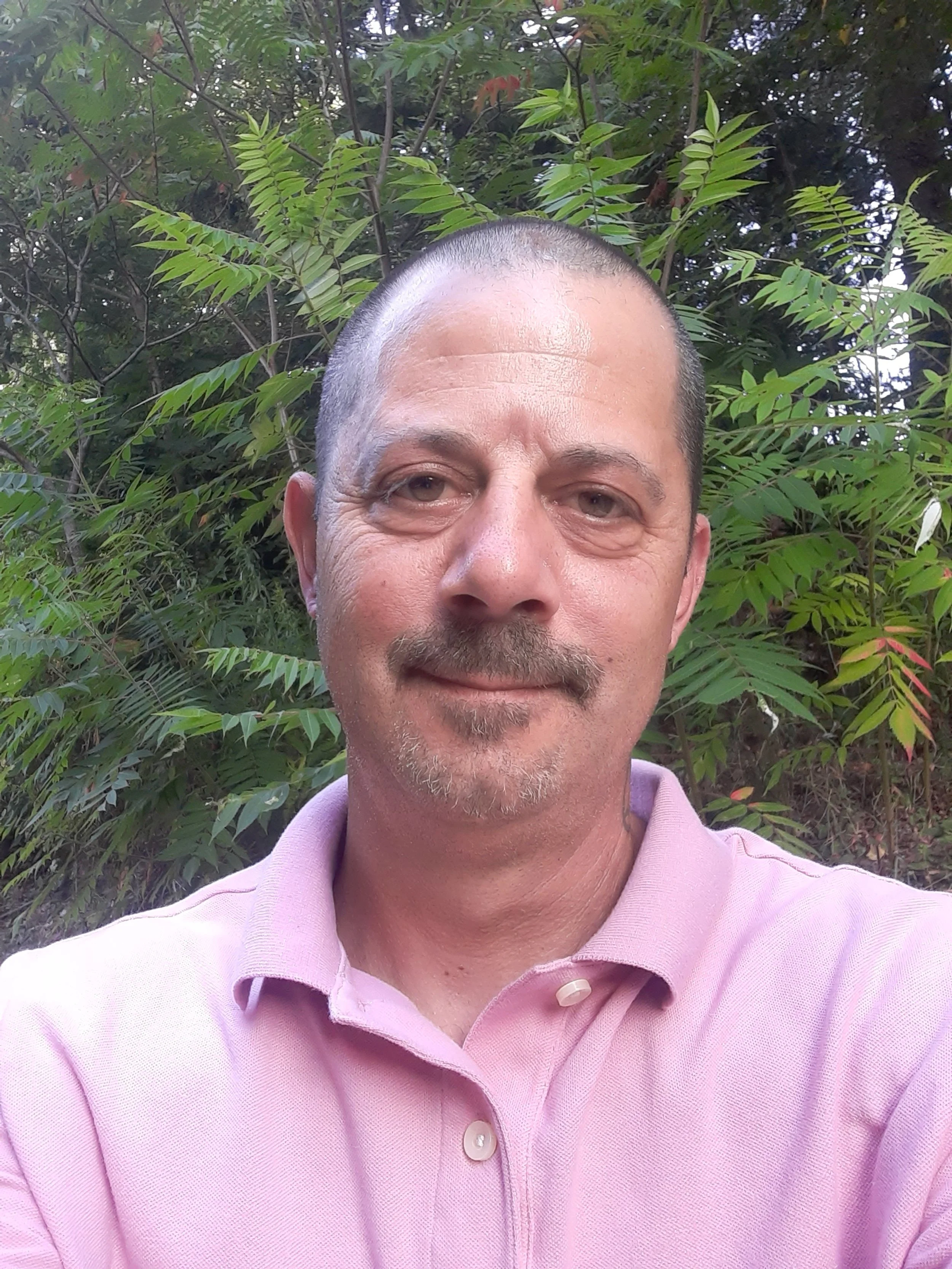 A man with short hair, a goatee, and a mustache smiling in front of green leafy trees, wearing a light pink polo shirt in an outdoor setting.