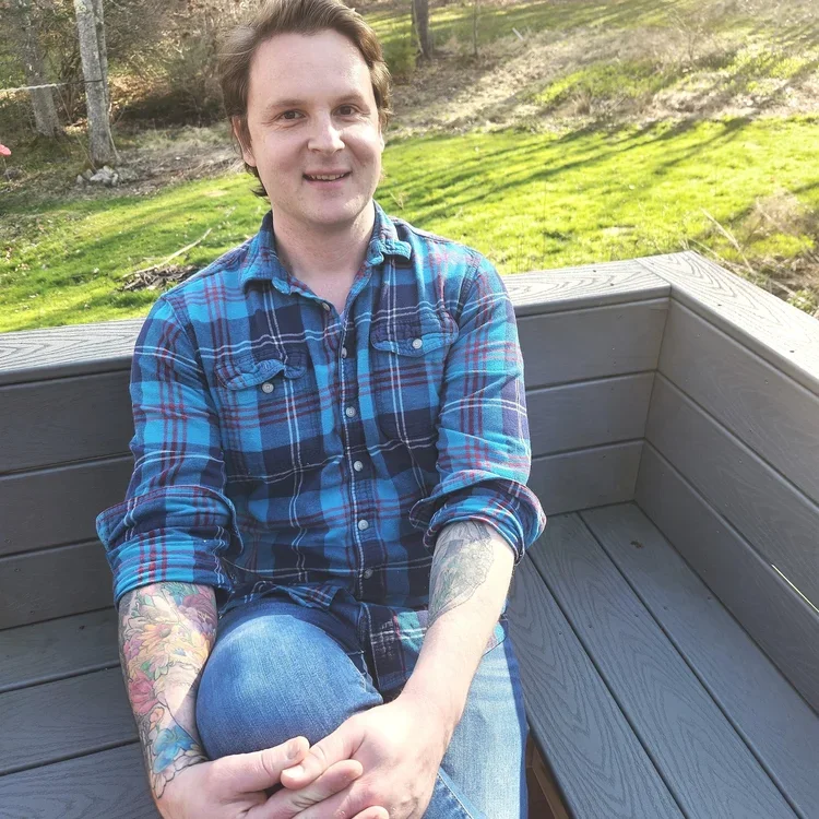 A young man with light skin, dark hair, and tattoos on his arms sitting on a wooden bench outdoors. He is smiling and wearing a blue plaid shirt and jeans, with the background of green grass and trees.