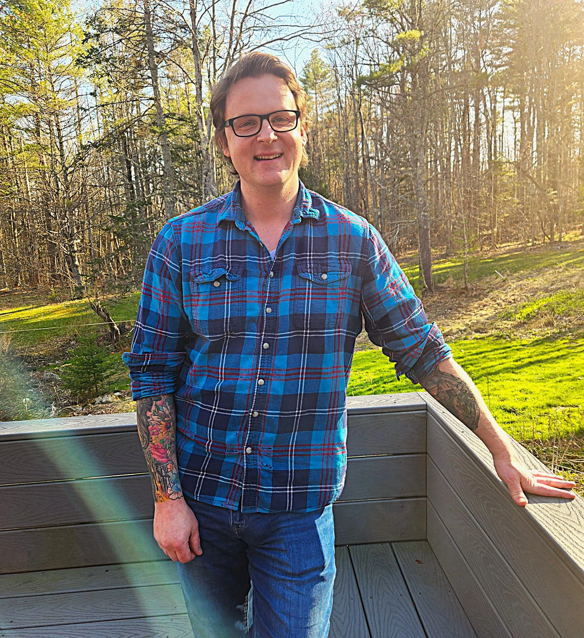 A young man with glasses and tattoos on his arms, wearing a blue plaid shirt and jeans, standing outdoors on a wooden deck with a forested background during sunset.