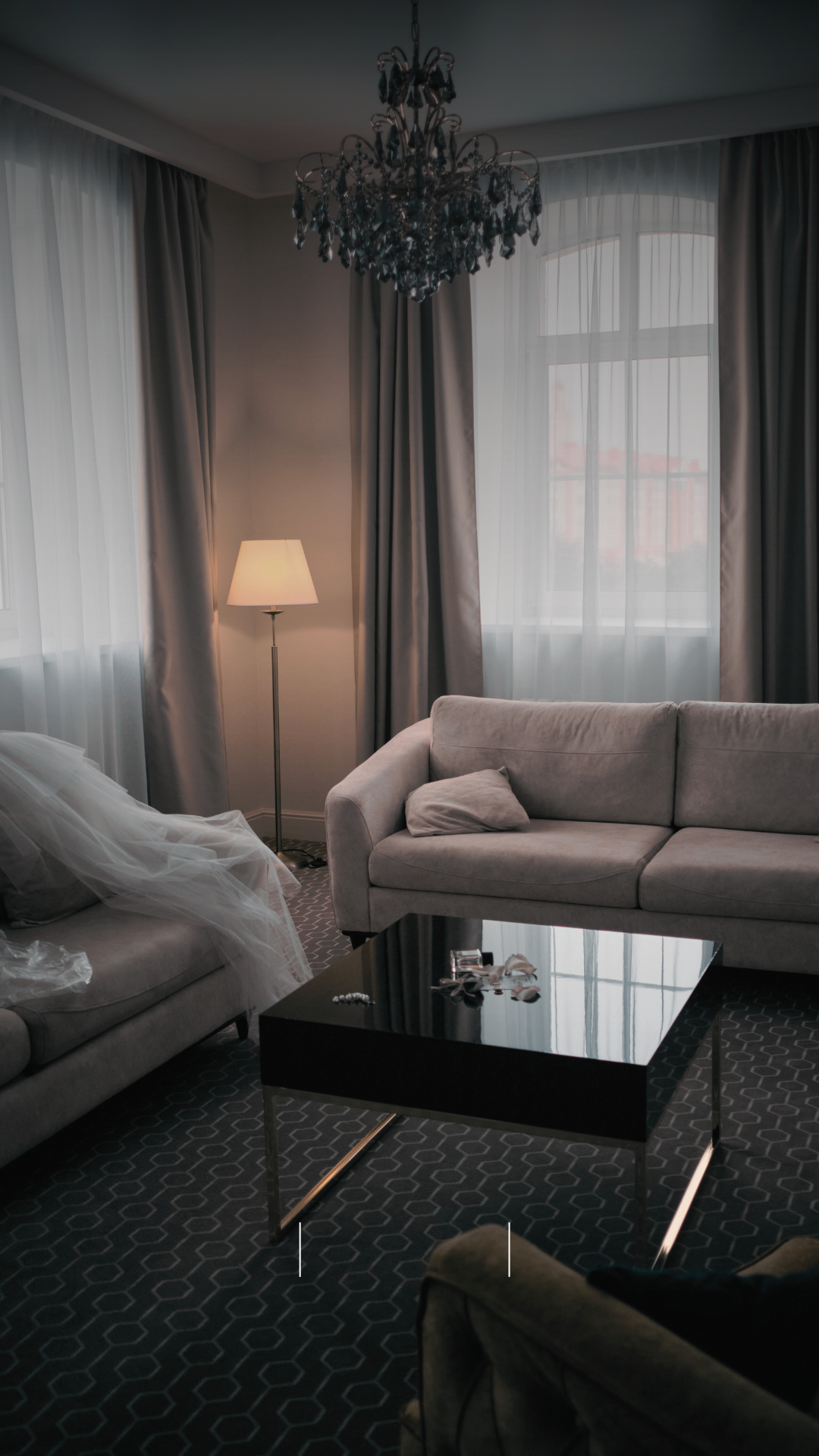 A living room with a chandelier, a floor lamp, beige sofas, a black coffee table, curtains, and a window letting in natural light.