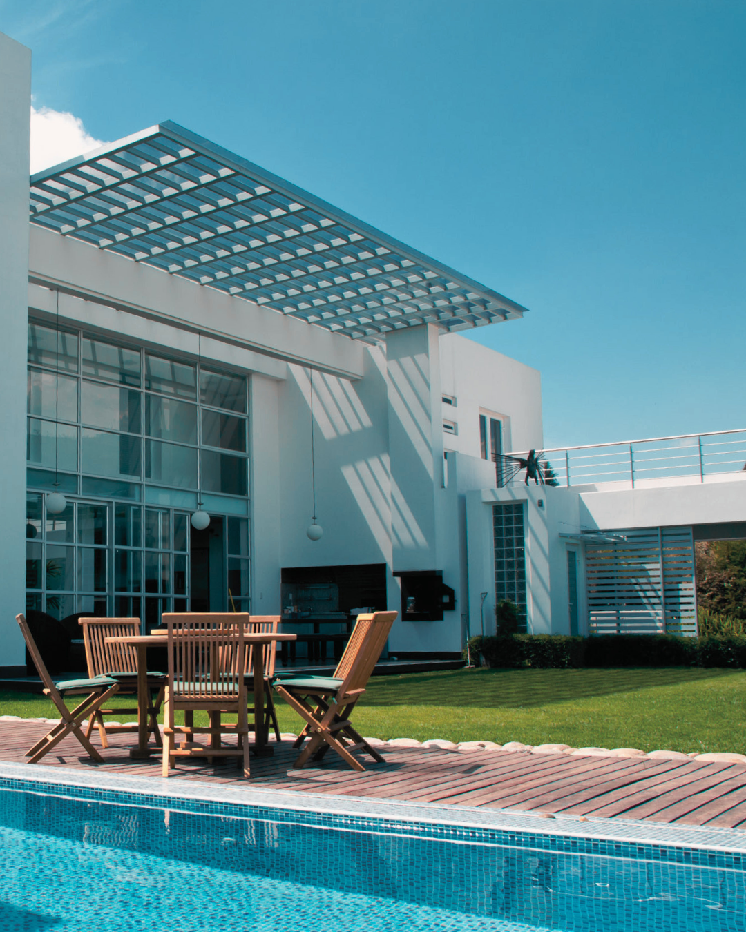 Modern house with large glass windows, outdoor pool, wooden table with chairs, balcony, and clear blue sky.