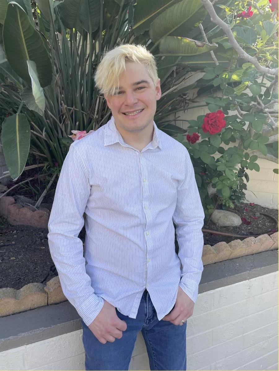 A blond man smiles in a garden of roses.
