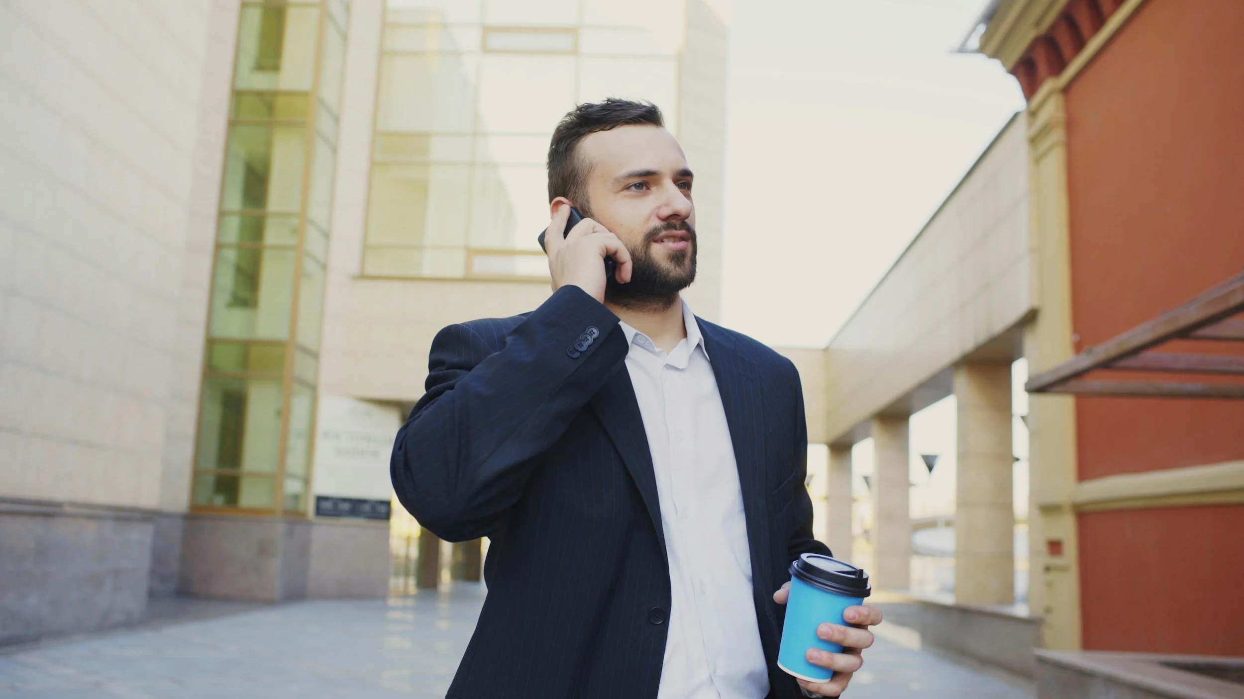 Businessman in a suit talking on a mobile phone while holding a coffee cup outside a modern building.