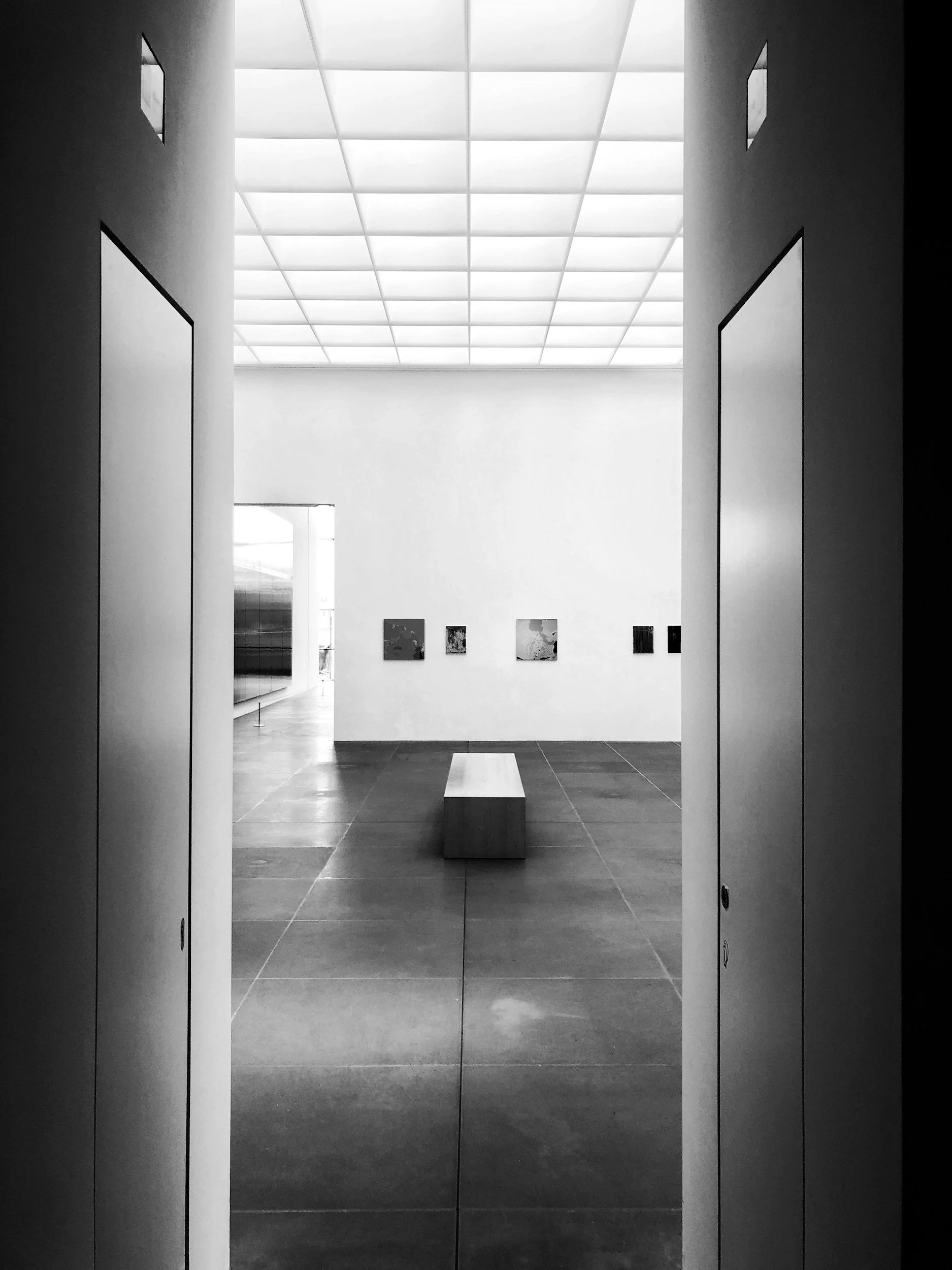 Black and white photo of an art gallery viewed through an open doorway. The gallery features a row of small artworks on the far wall, a long bench in the center, and gallery lighting on a grid ceiling.