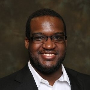 A smiling man with glasses, wearing a black suit and white shirt, against a dark background.